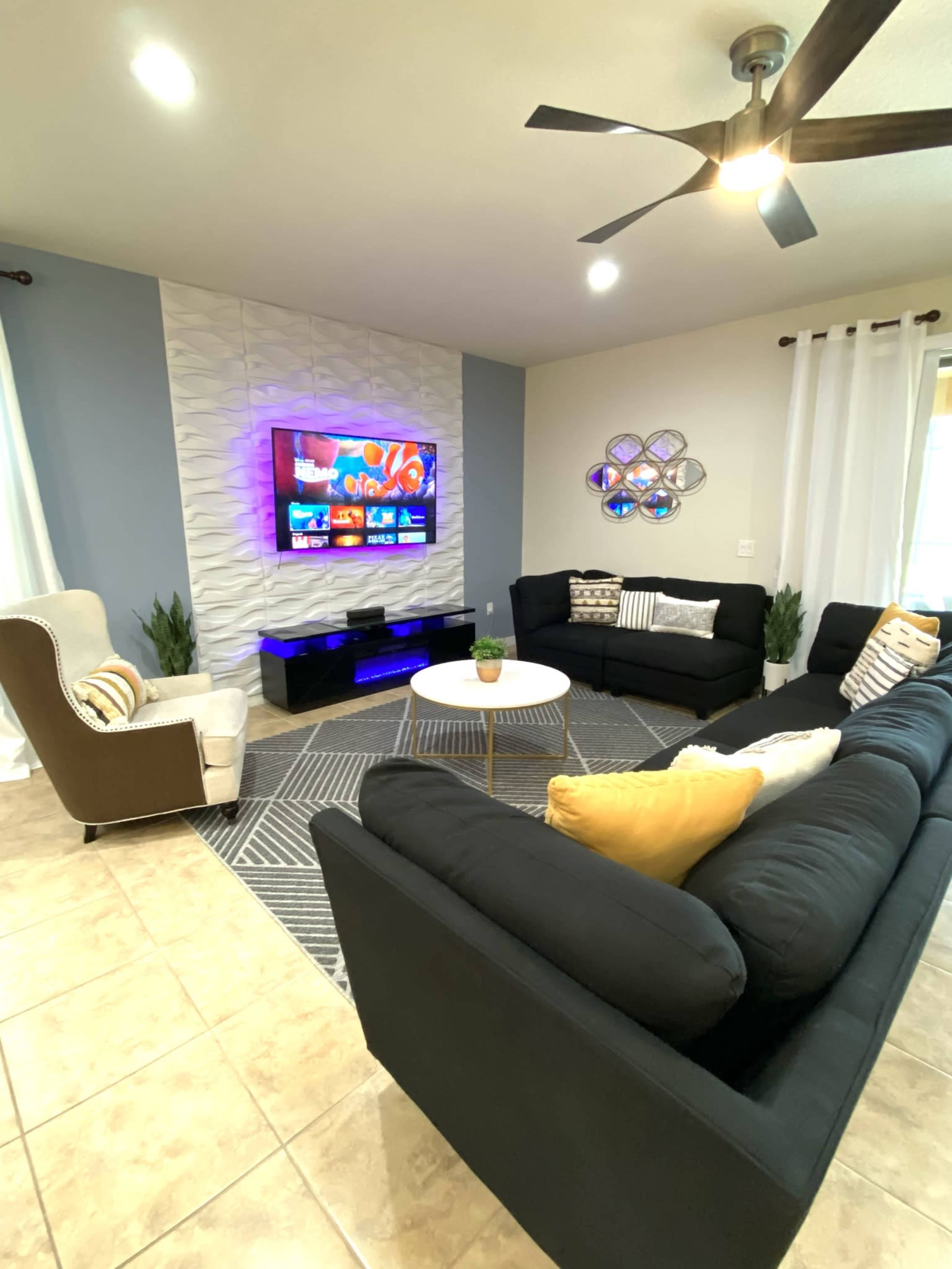 A living room features a sectional sofa, an armchair, a round coffee table, a television mounted on a textured wall, and decorative plants.