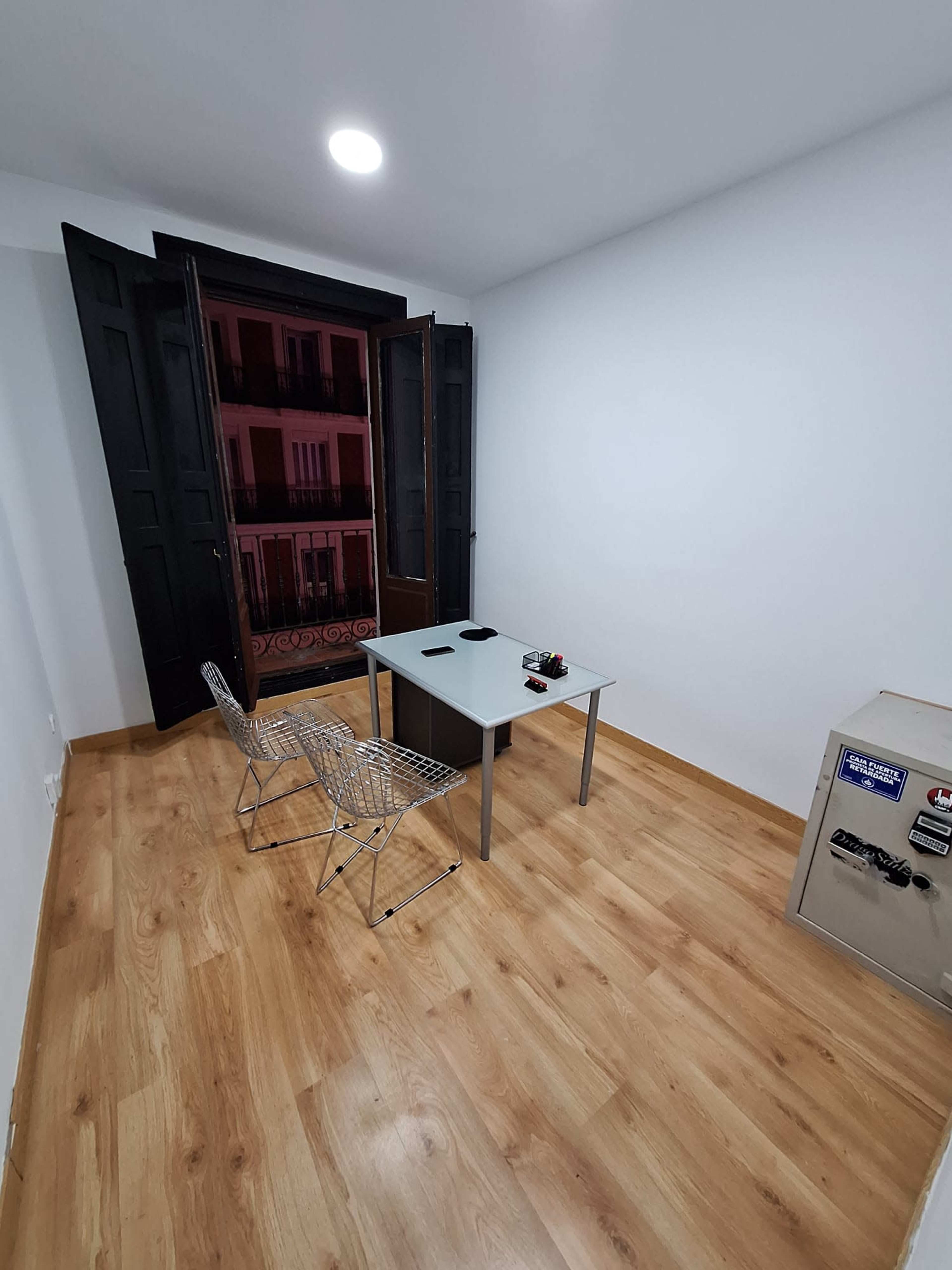 The image shows a small office space with a glass table, two wire chairs, and a safe, all in front of open double windows that reveal a view of a building's exterior.