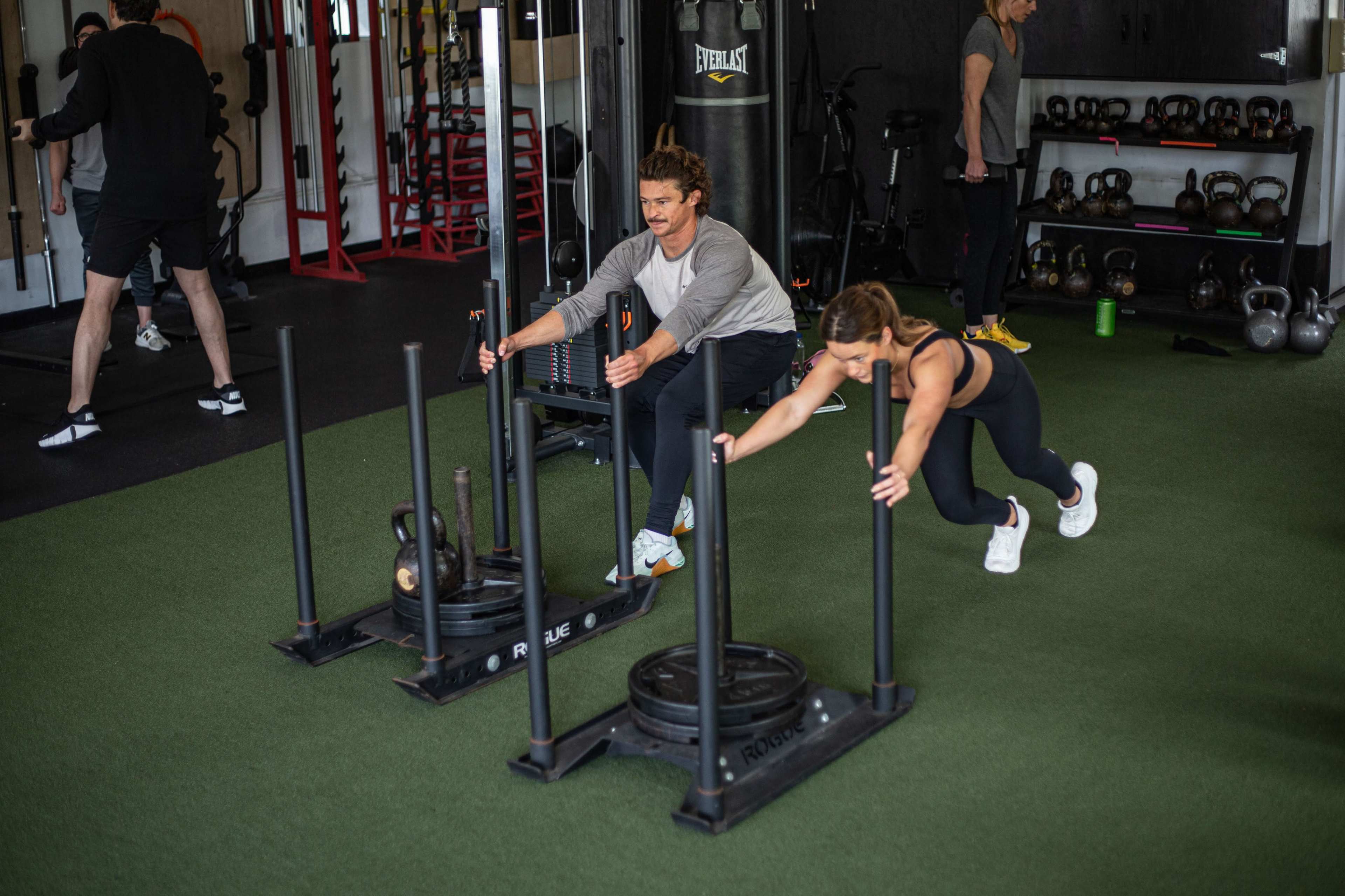 Two individuals are pushing sleds across a gym floor with various weights in a fitness training setting.