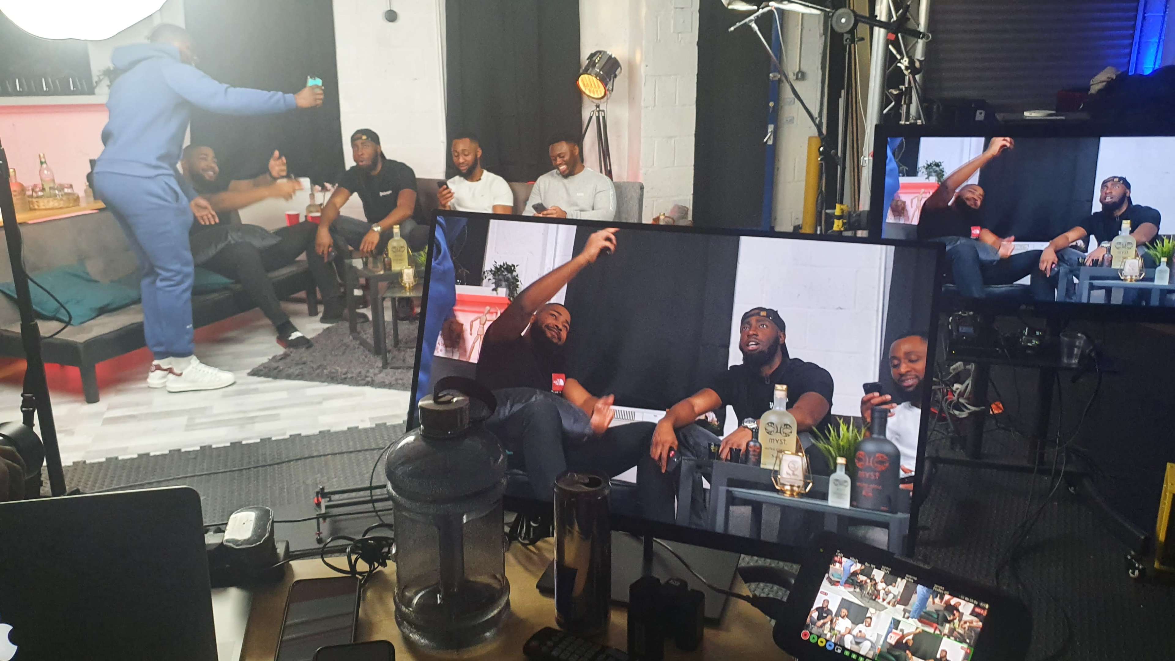 A group of men are sitting on a couch in a studio, while one man stands and talks, capturing the scene on a camera display in the foreground.