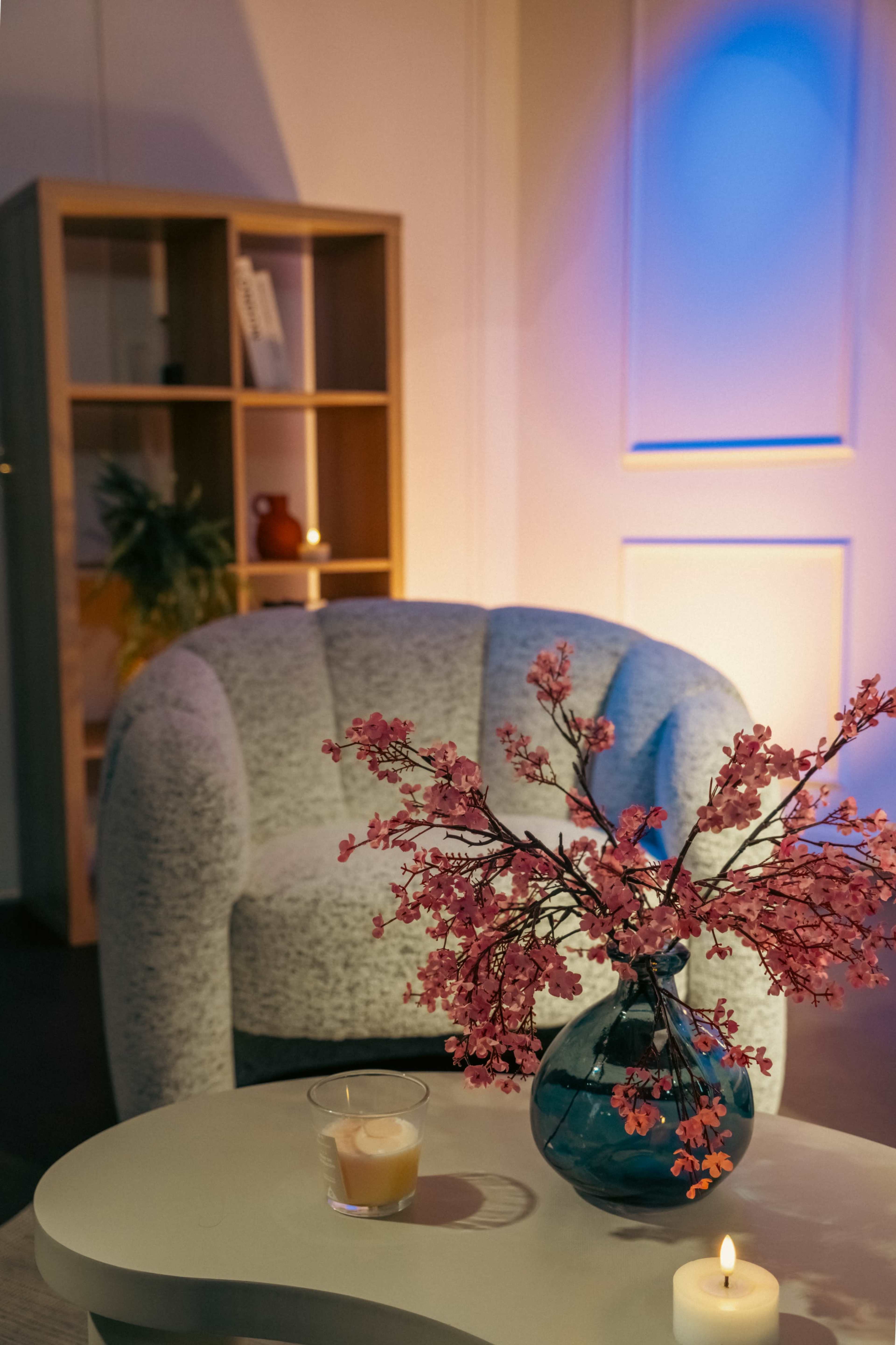 A cozy interior scene features a plush armchair beside a table with a decorated vase filled with pink branches, a candle, and a drink.
