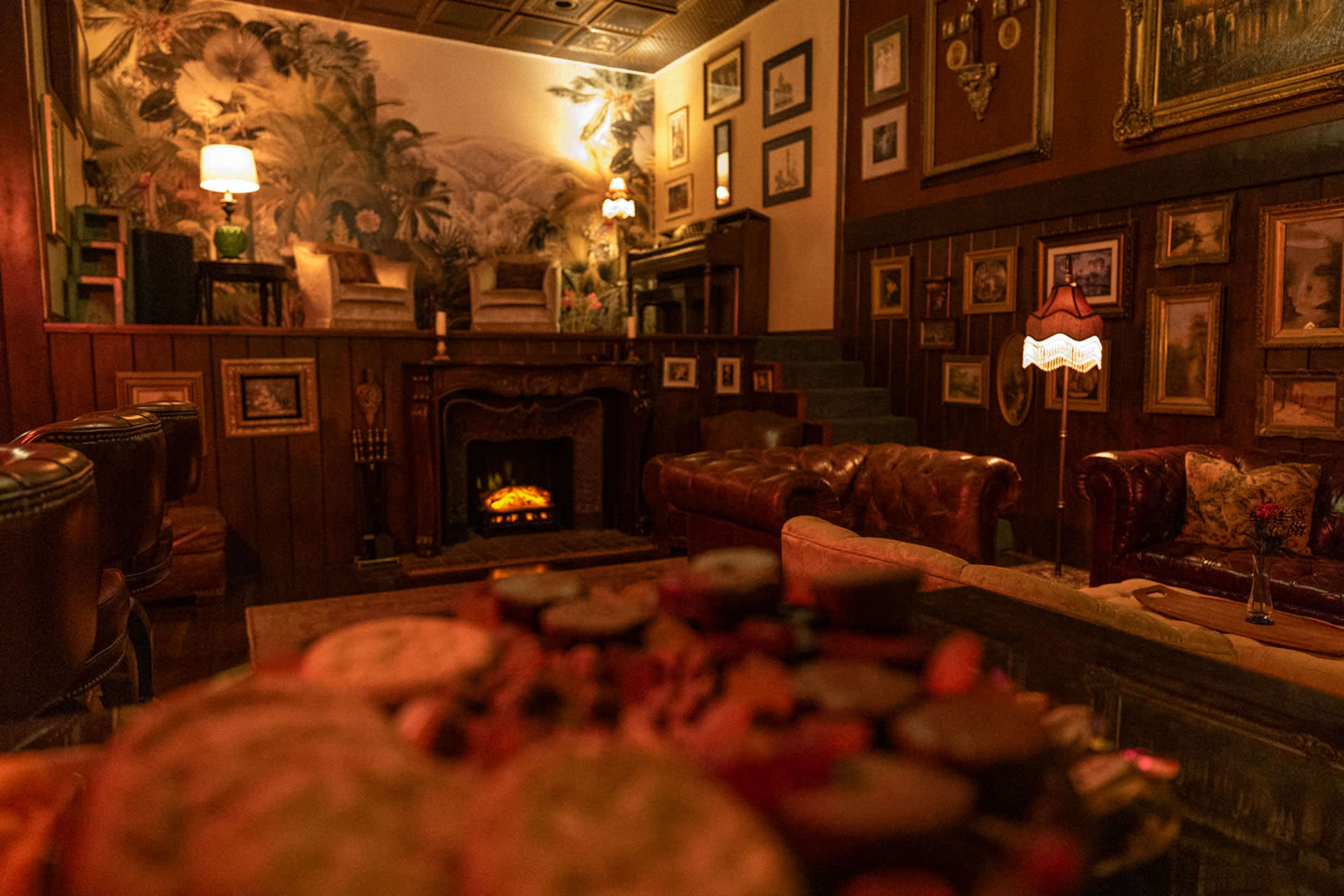 The image shows a cozy lounge area with a fireplace, leather seating, and wall decor featuring framed pictures, illuminated by soft lighting.