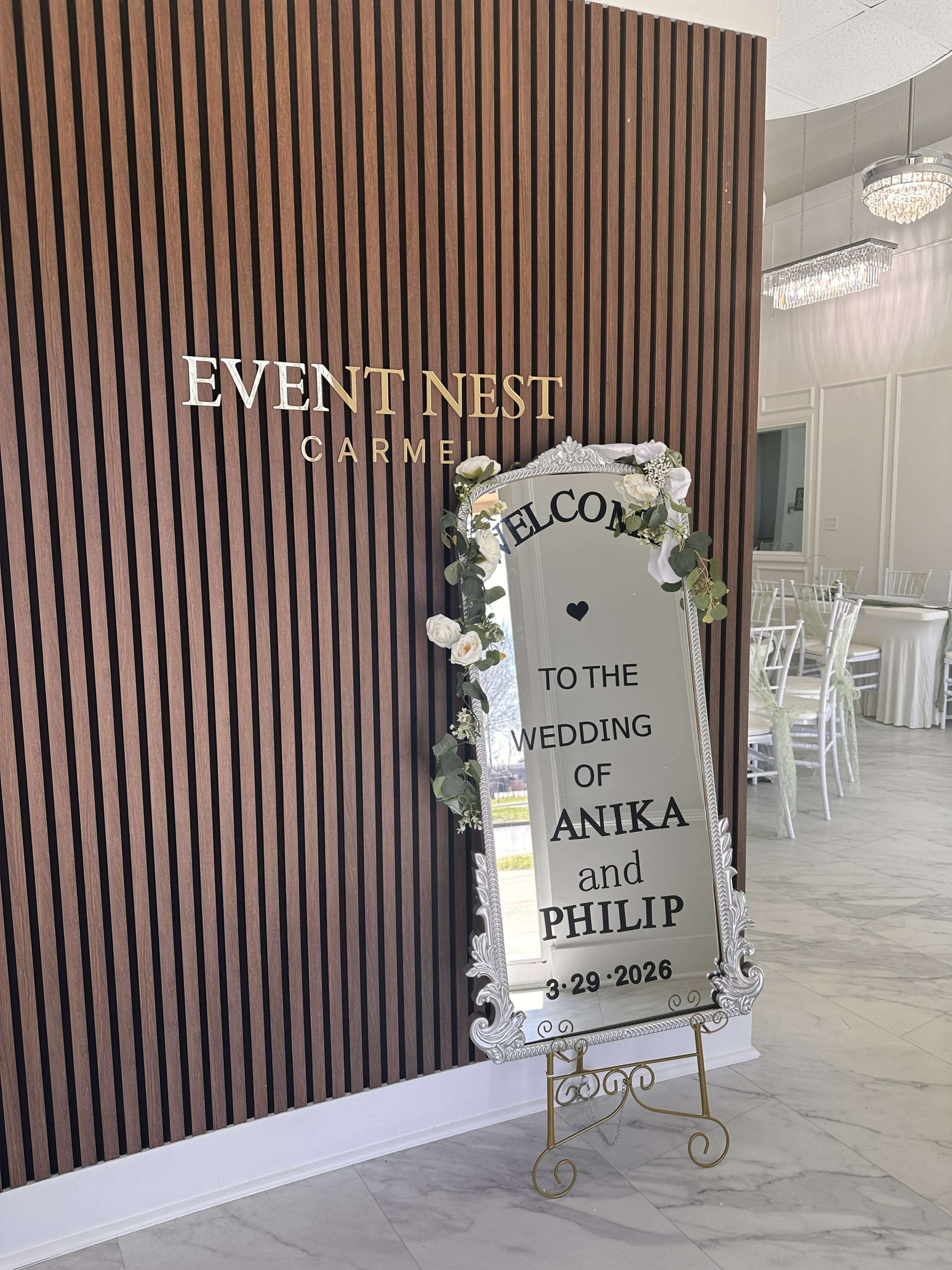 A decorative mirror displaying a welcome message for Anika and Philip's wedding is positioned against a wooden-paneled wall in the Event Nest venue.