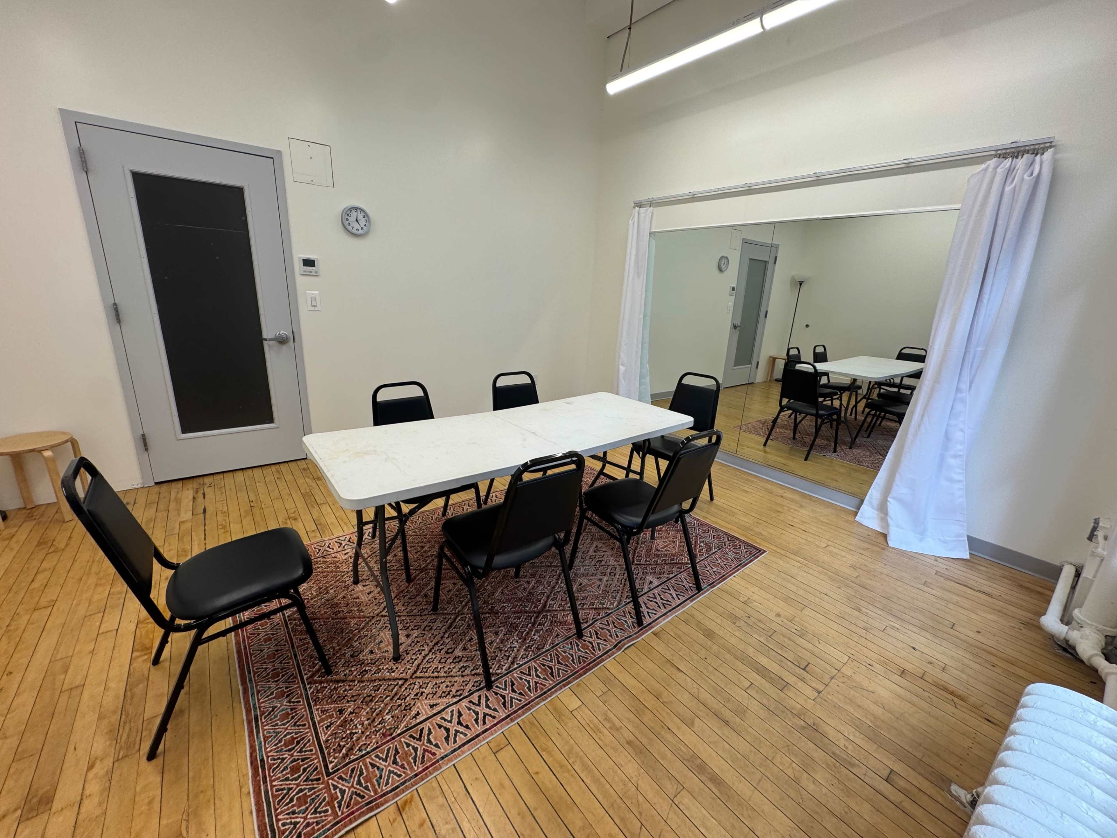 A simple meeting room features a rectangular table surrounded by black chairs, with a mirrored wall and a clock visible.