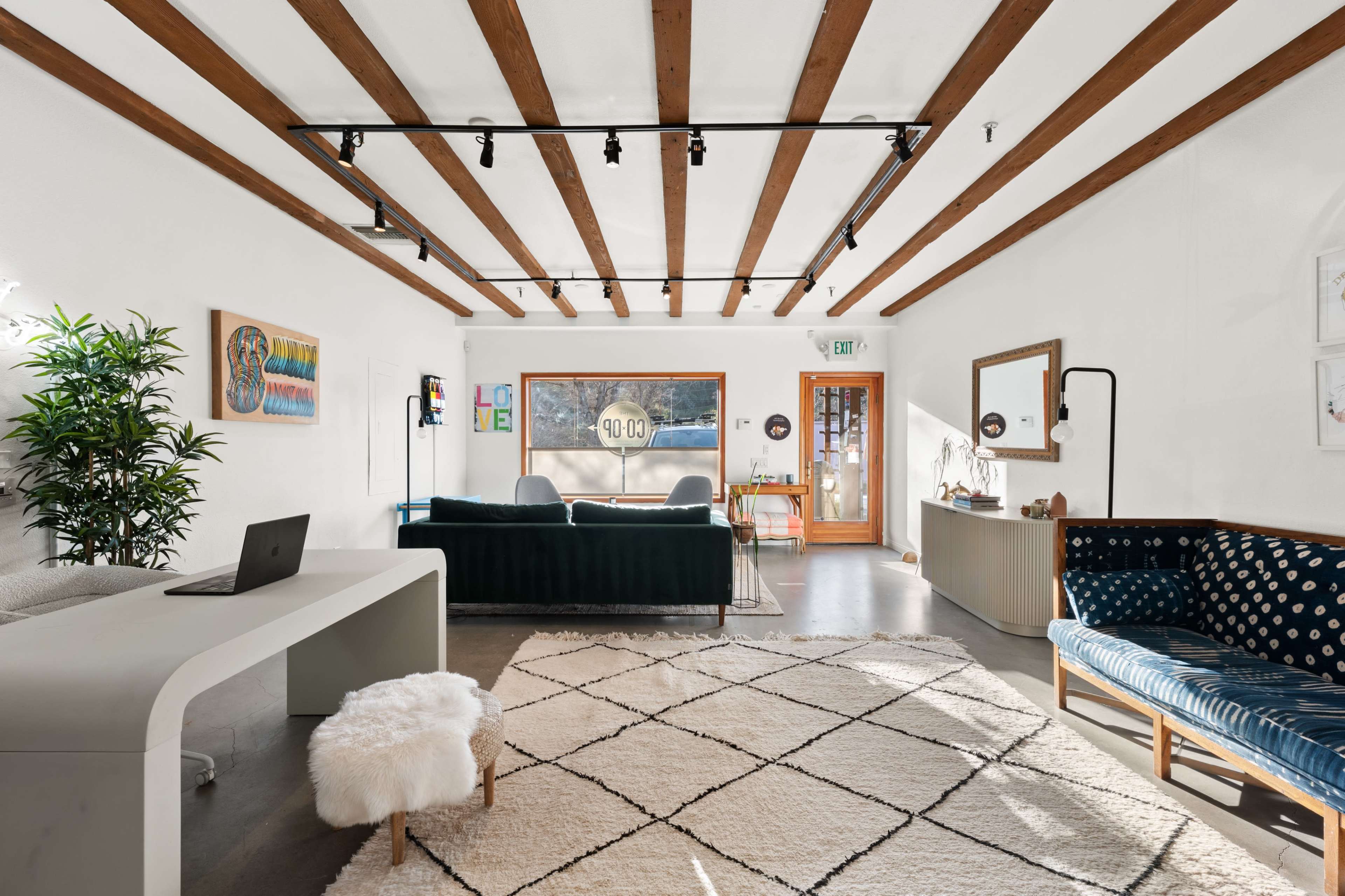 A modern interior space with exposed wooden beams, a large area rug, a desk with a laptop, green and blue seating areas, and a front entrance with a large window.