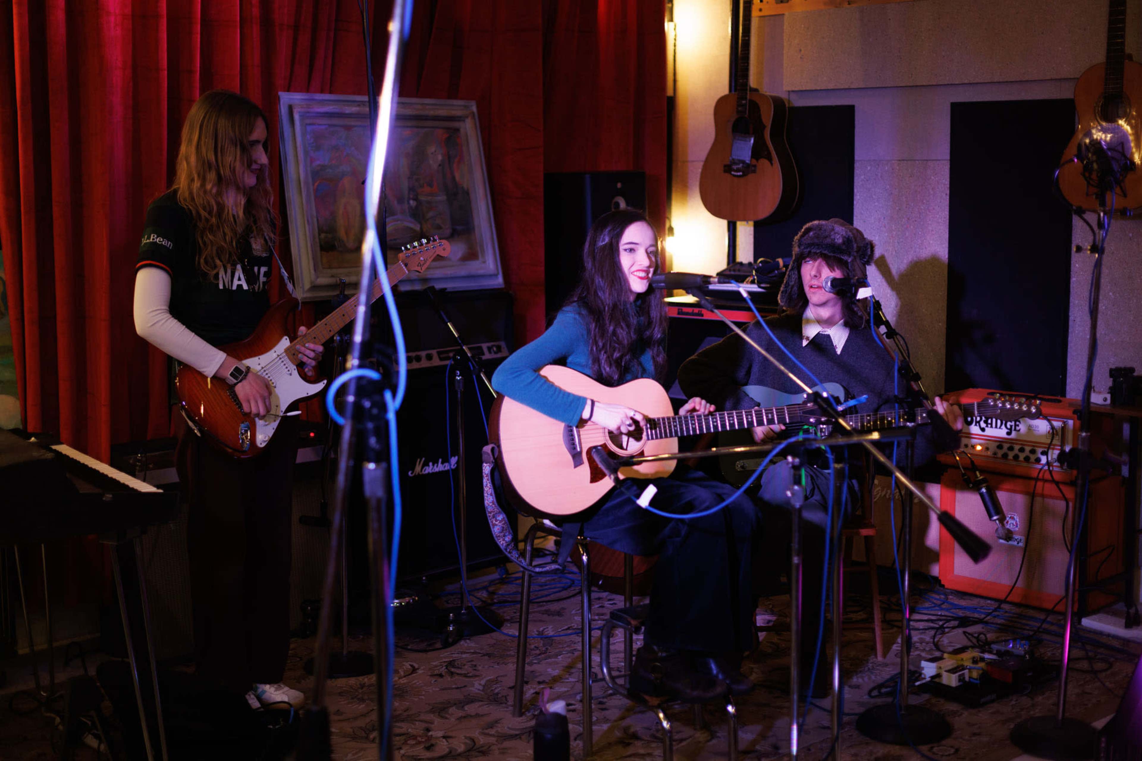 Three musicians perform in a cozy studio setting, with a red backdrop and various musical instruments visible.