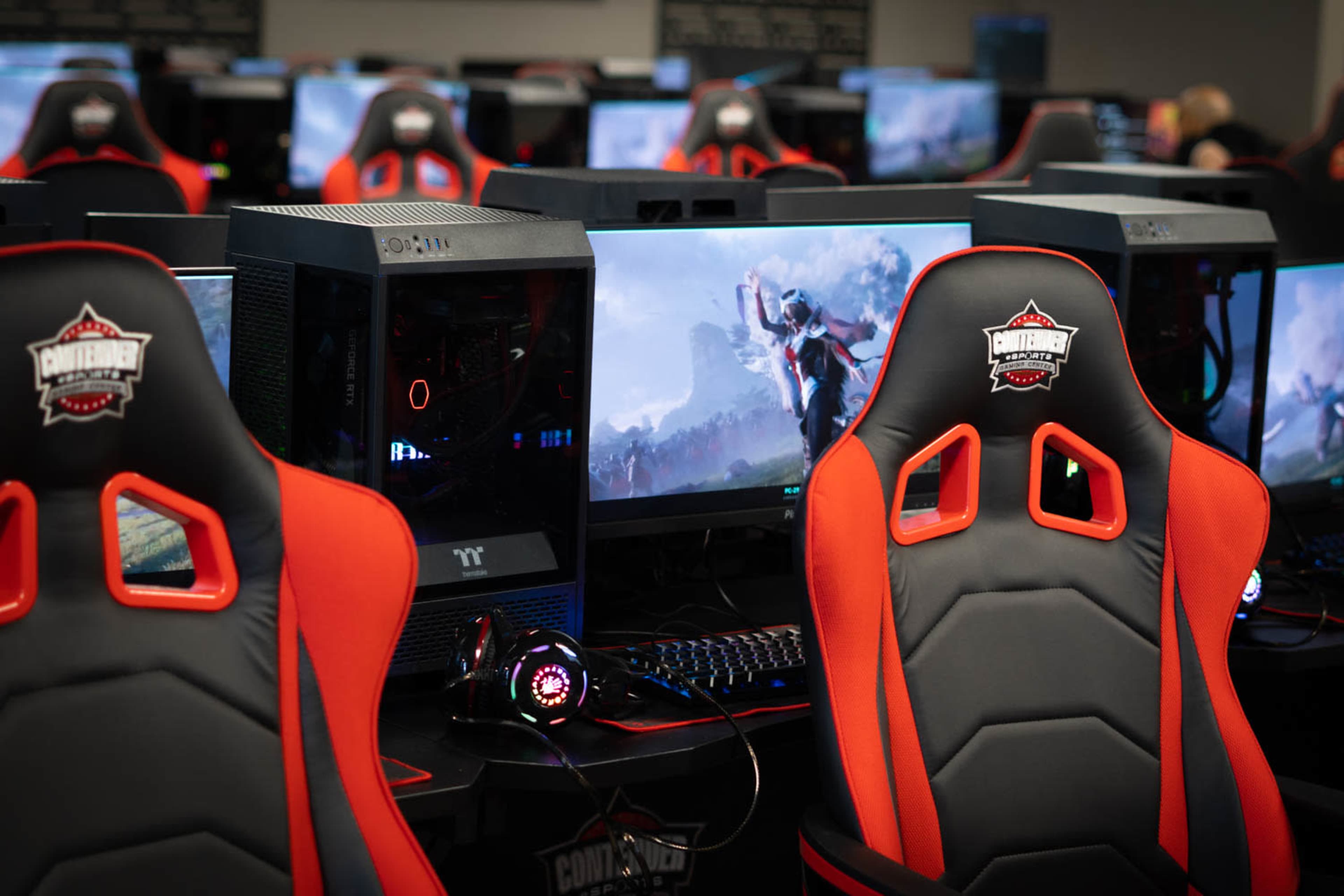 A row of computer gaming stations with high-backed racing-style chairs and monitors displaying a video game scene.
