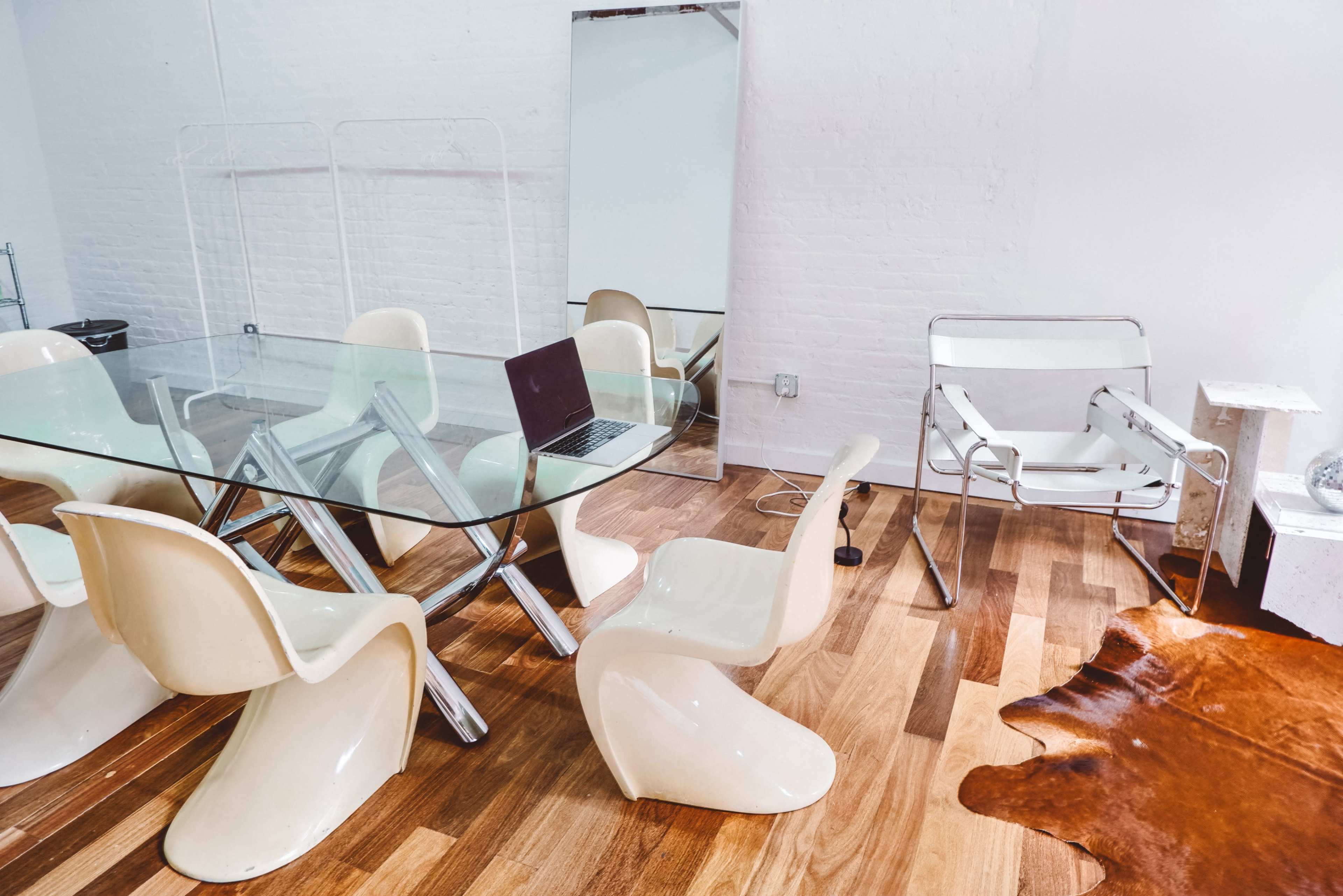 A modern office space with a glass dining table, several white plastic chairs, a laptop on the table, and a cowhide rug on the wooden floor.