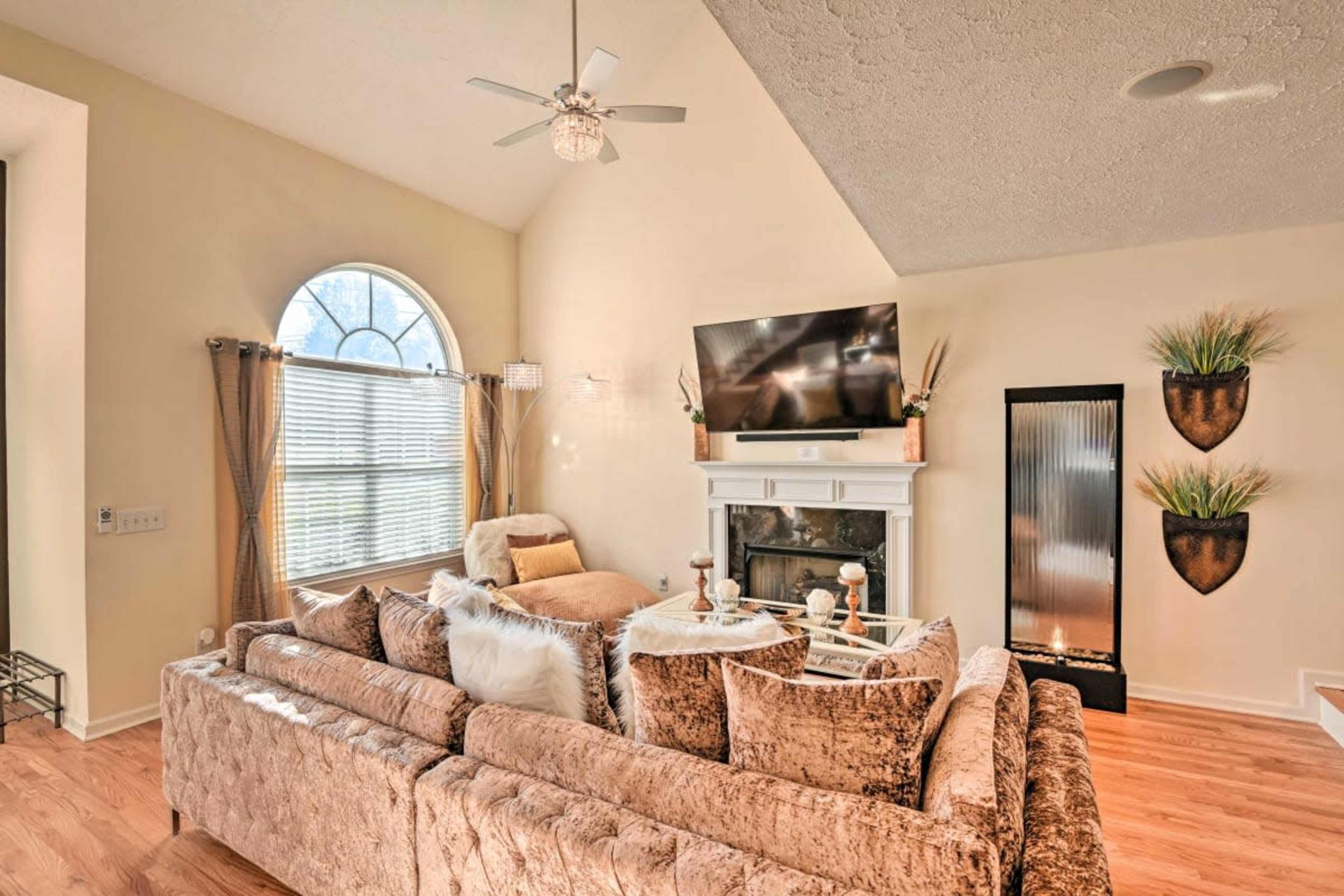 The living room features a beige sectional sofa, a wall-mounted television, and a decorative fireplace under a high, sloped ceiling.