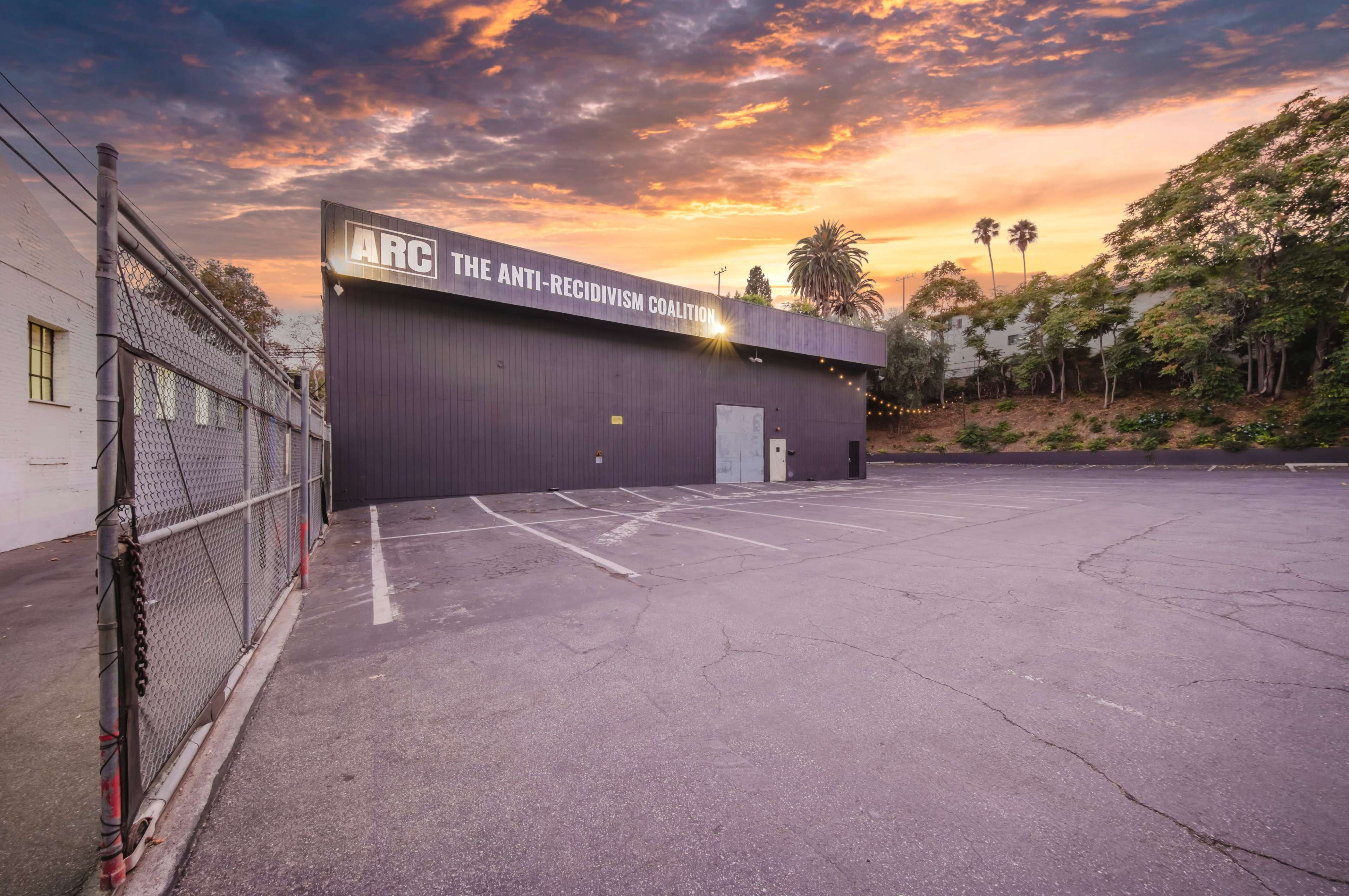 The image shows a large, modern building with a sign reading "ARC THE ANTI-RECIDIVISM COALITION" against a colorful sunset sky.