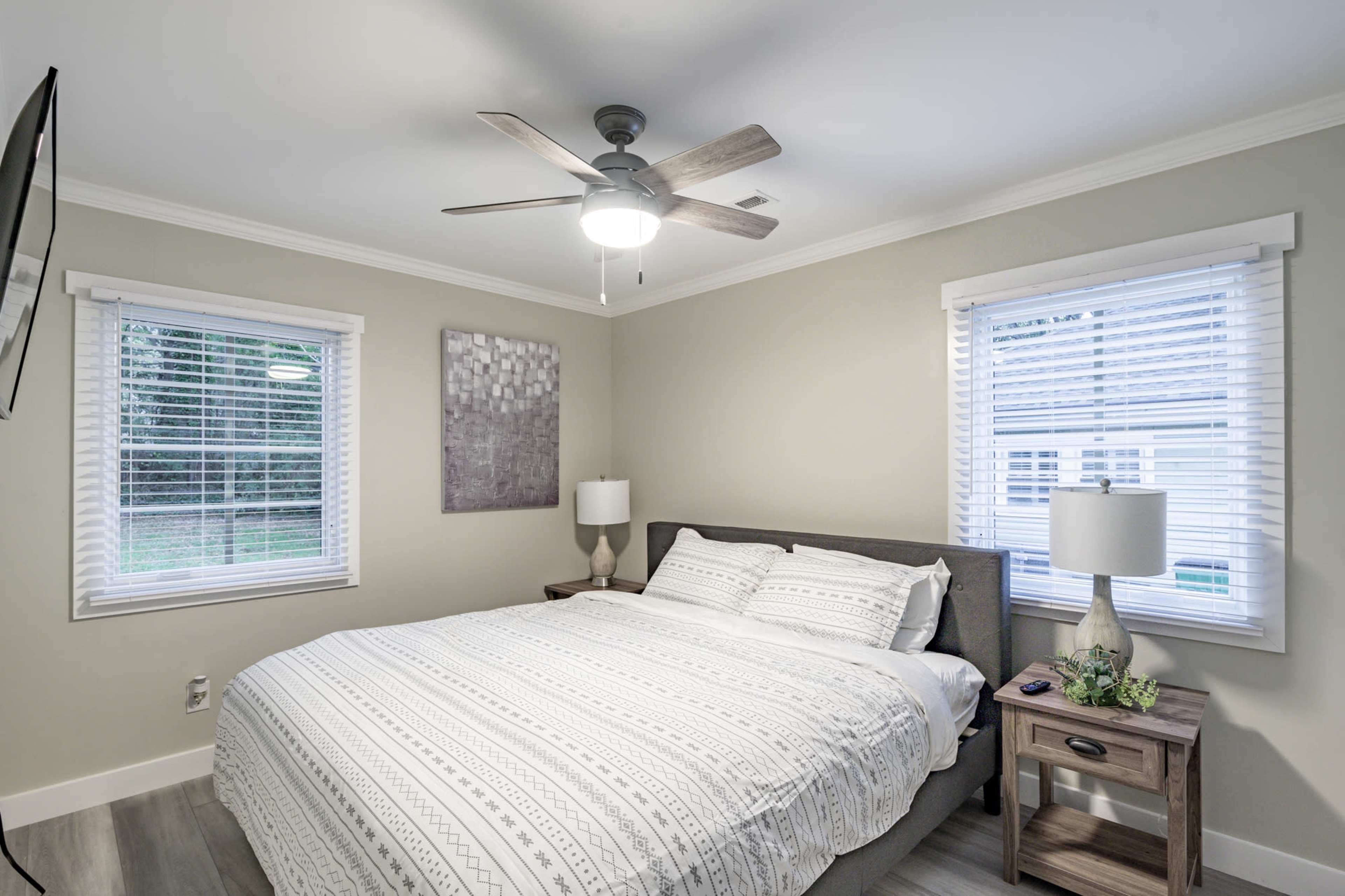 The image shows a neatly arranged bedroom featuring a large bed with a patterned duvet, two windows with blinds, and a nightstand with a lamp.