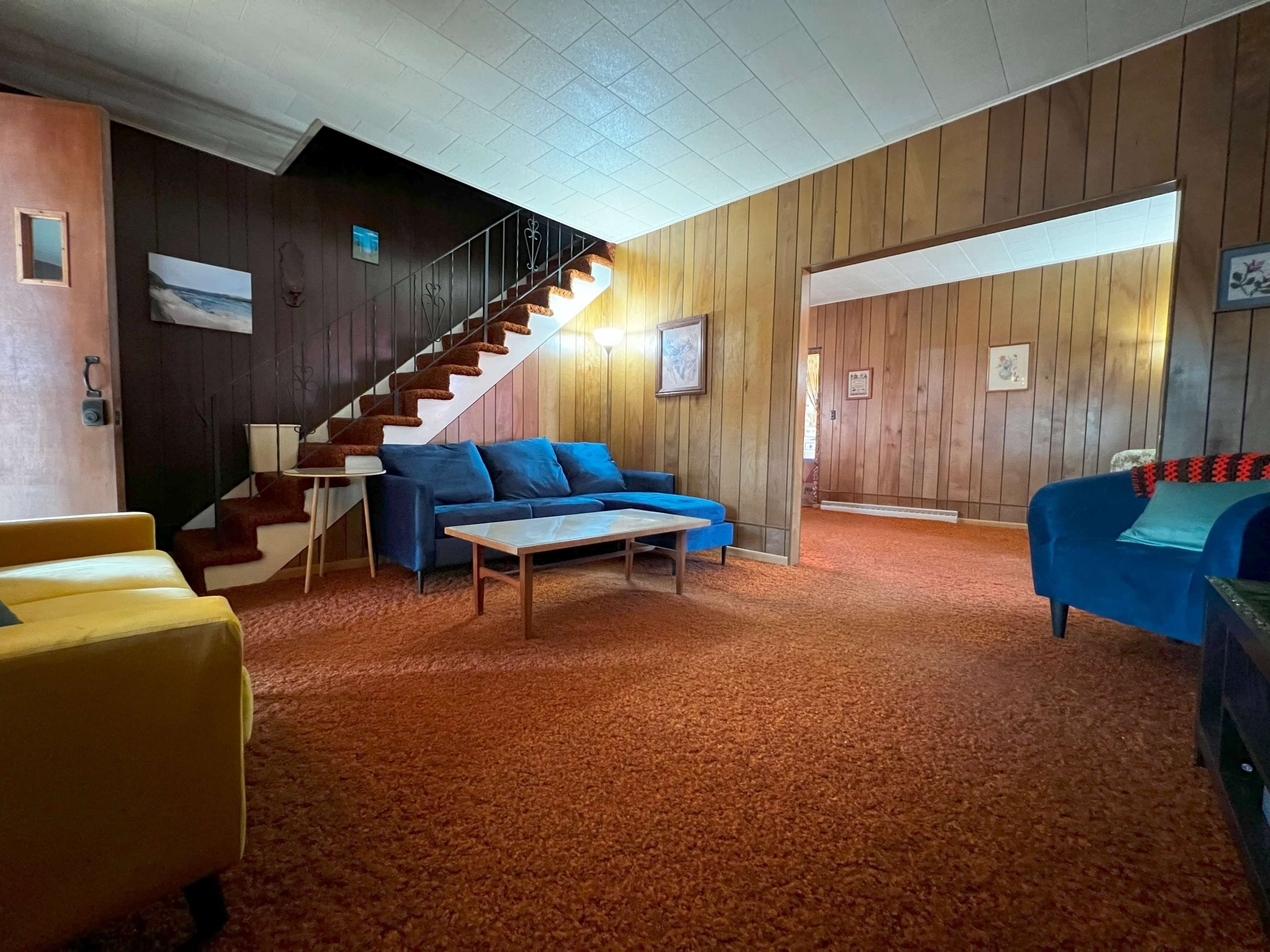 A living room features blue sofas, a wooden staircase, and orange carpeting.