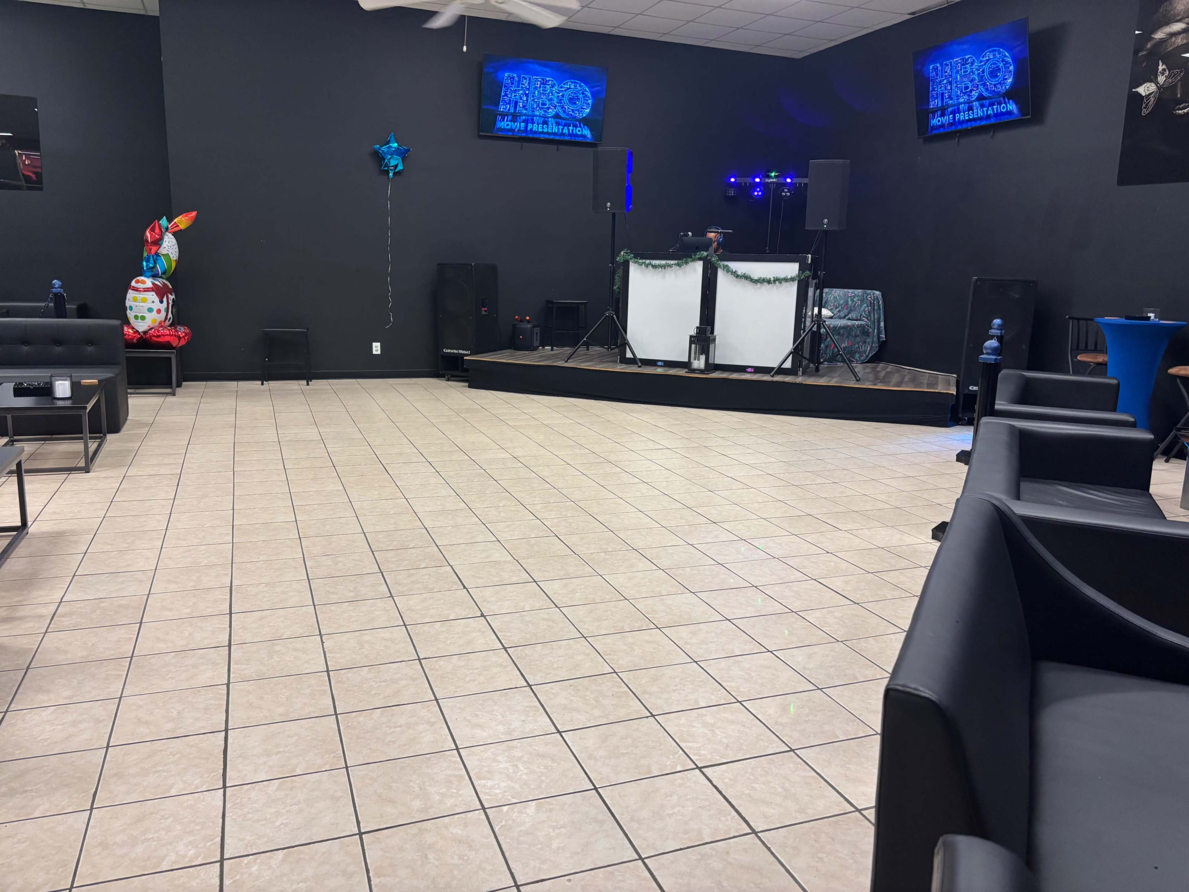 The image shows an indoor event space with tiled flooring, black walls, a stage set up for a performance, and seating areas consisting of black sofas.