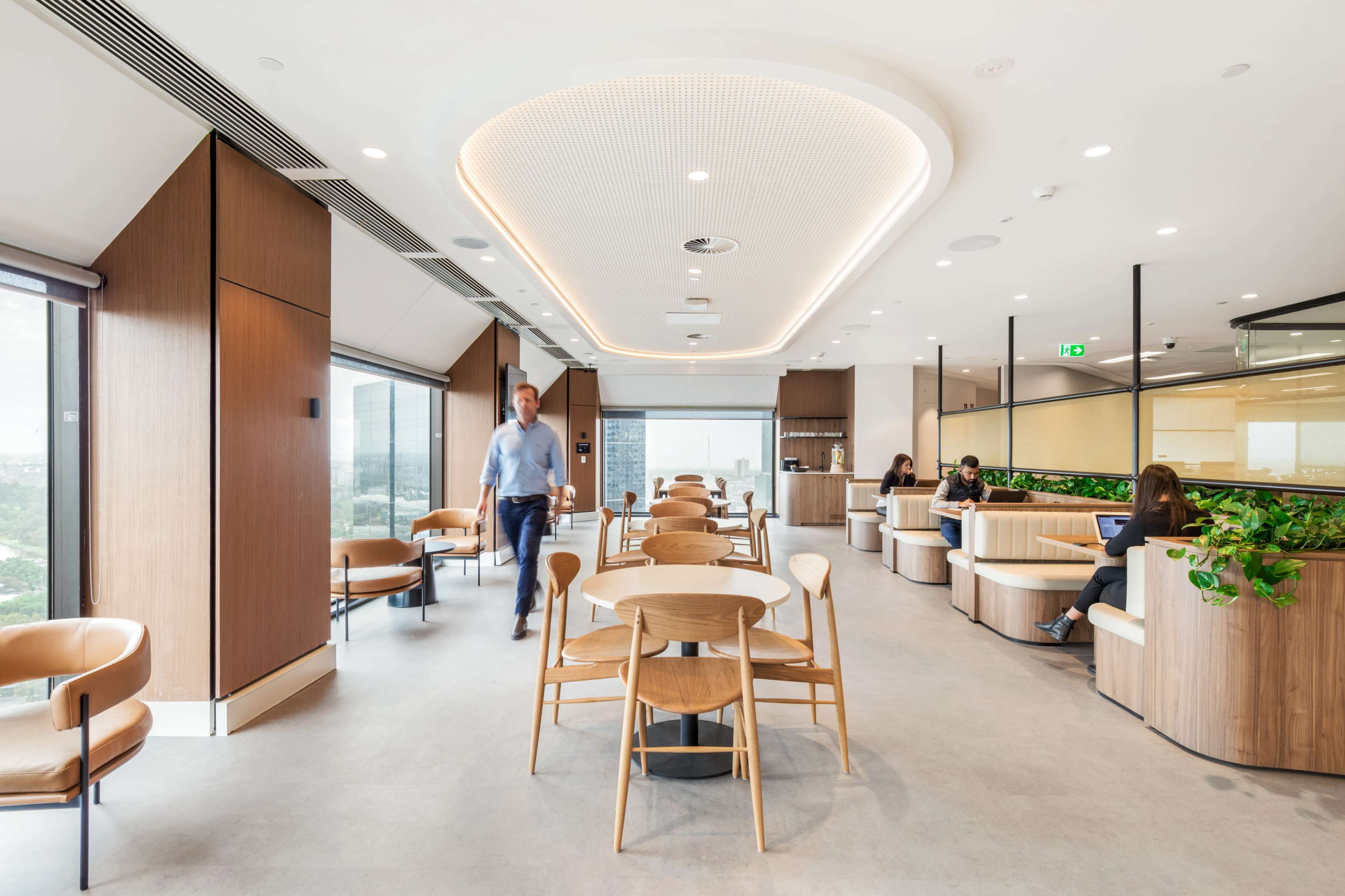 A modern office space features wooden tables and chairs, large windows with city views, and individuals working and relaxing in various areas.