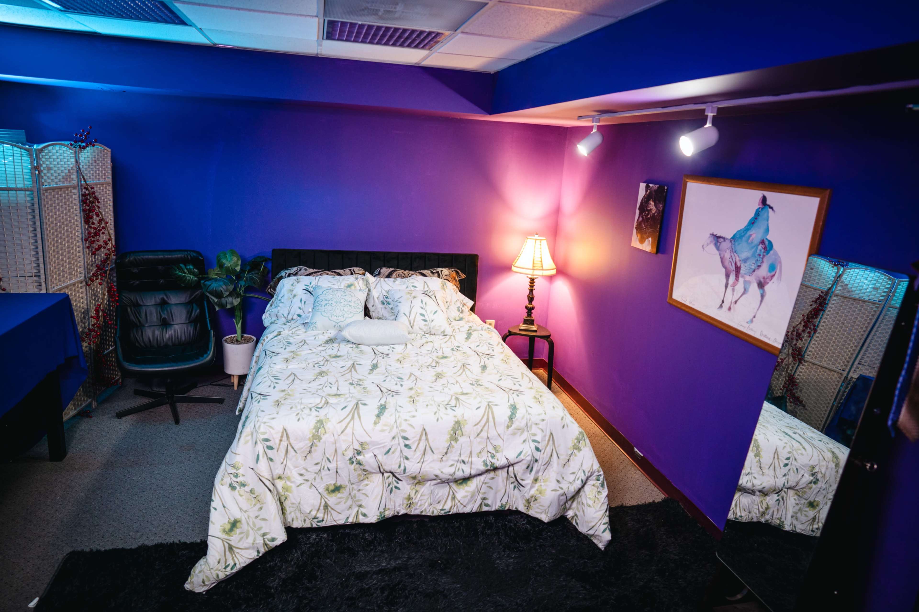 The image shows a bedroom with a purple accent wall, a king-size bed covered in a patterned comforter, a chair, and a large mirror.