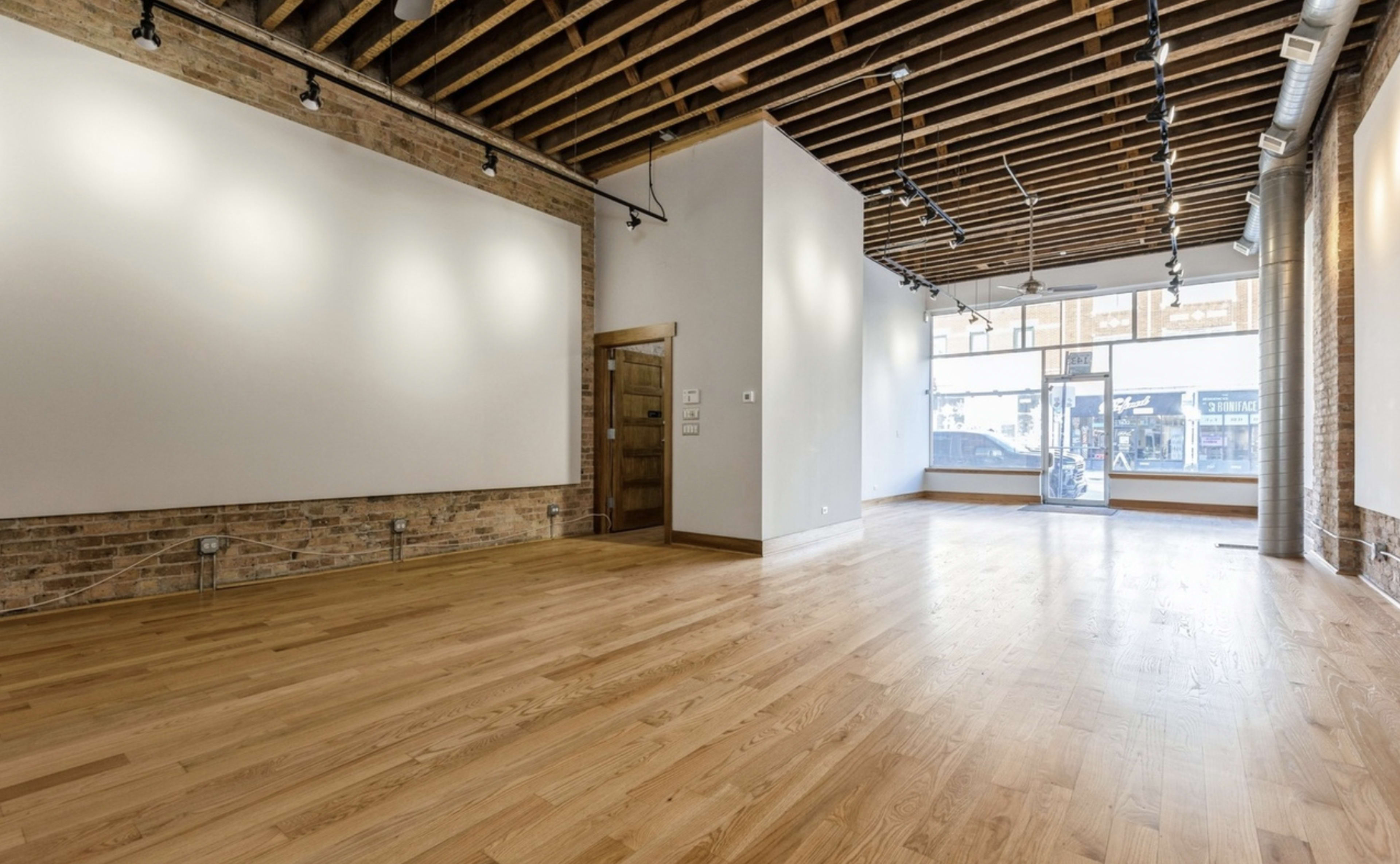 The image shows an empty commercial space with hardwood floors, exposed brick walls, and large windows allowing natural light to enter.