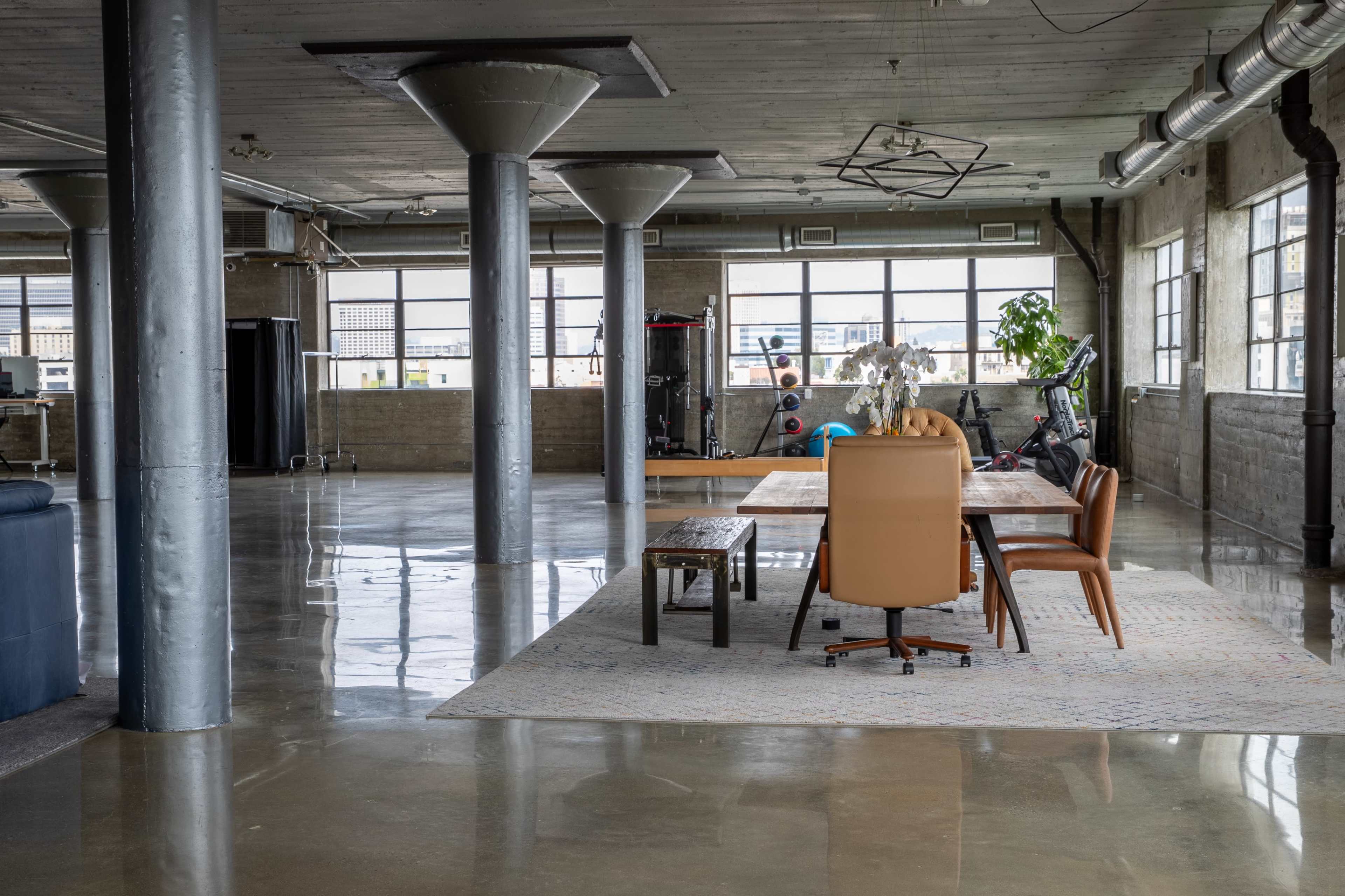 A spacious, modern industrial-style loft with polished concrete floors, a wooden dining table surrounded by chairs, and large windows allowing ample natural light.