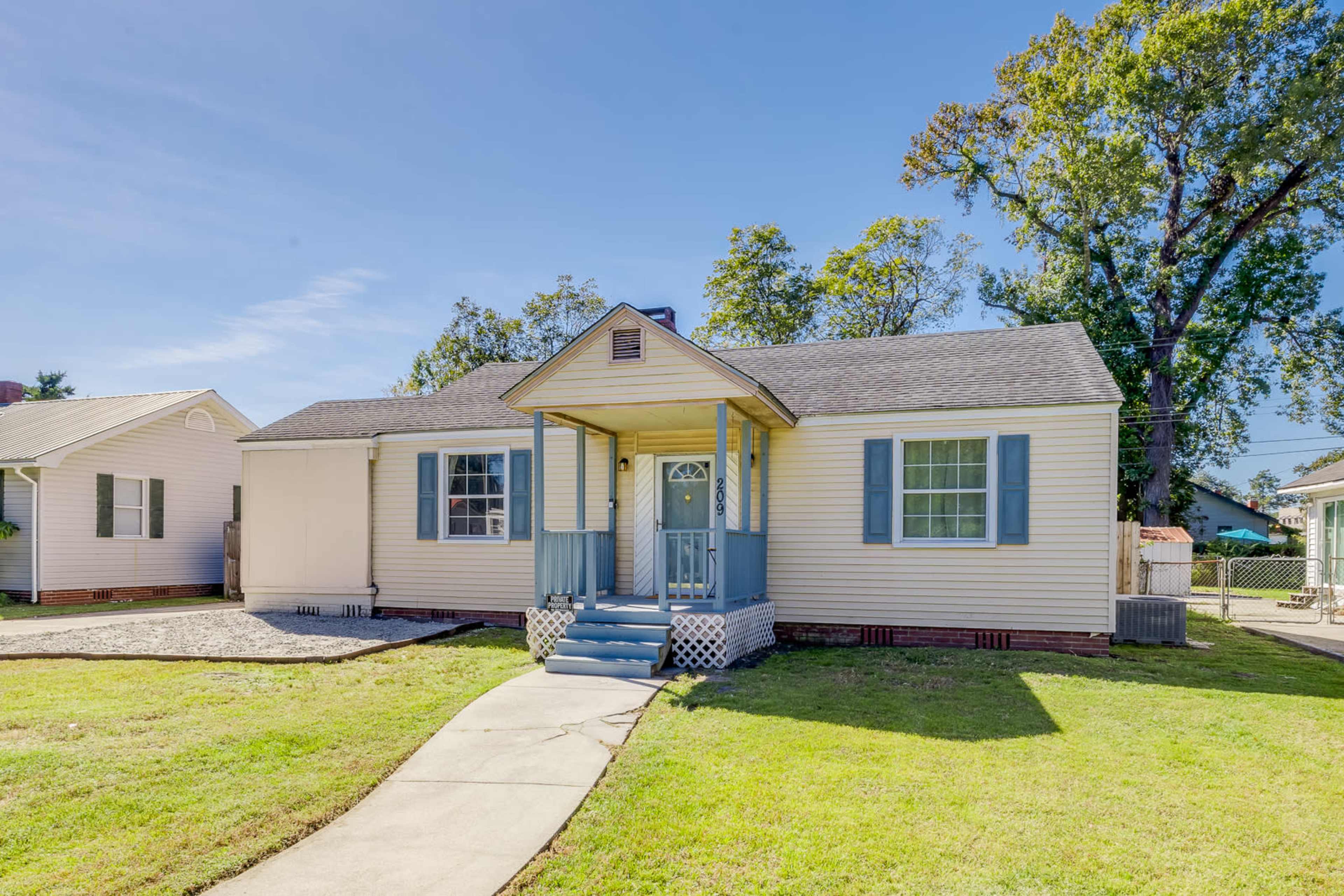The image shows a single-story, light-colored house with blue shutters and a front porch, surrounded by a lawn and nearby trees.