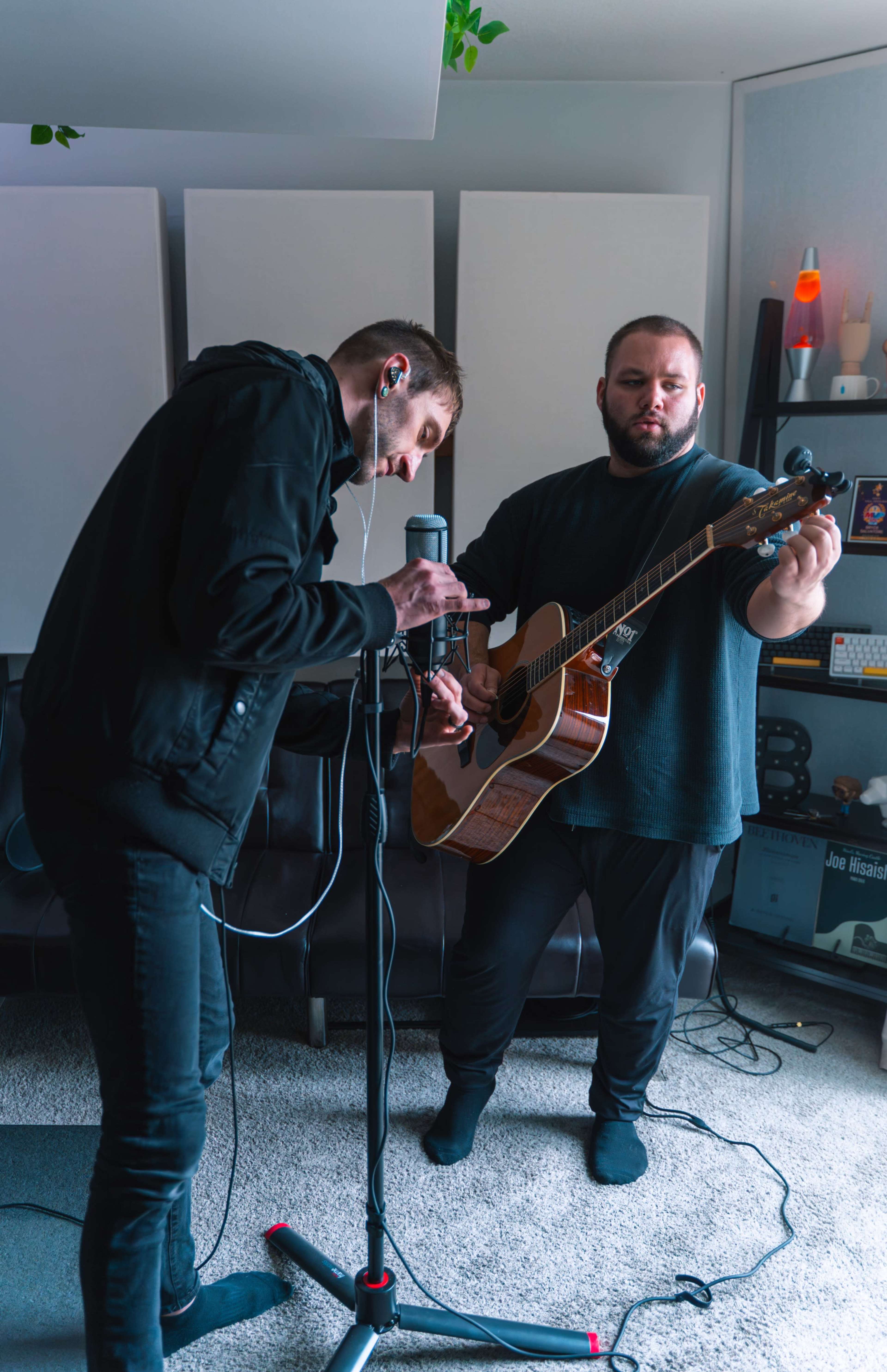 Two musicians are in a room, with one playing an acoustic guitar while the other adjusts a microphone and listens through earbuds.