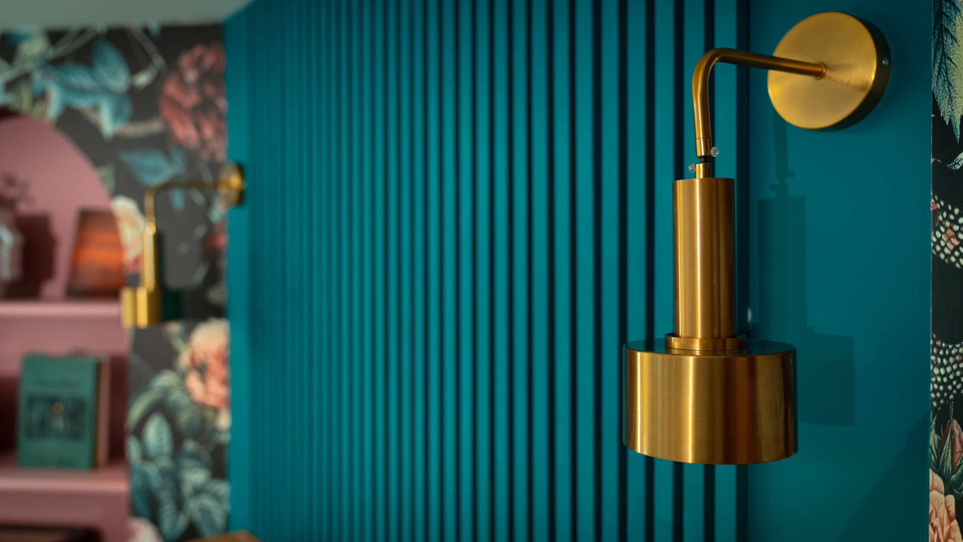 A brass wall sconce is mounted on a teal vertically striped wall, with floral wallpaper visible in the background.