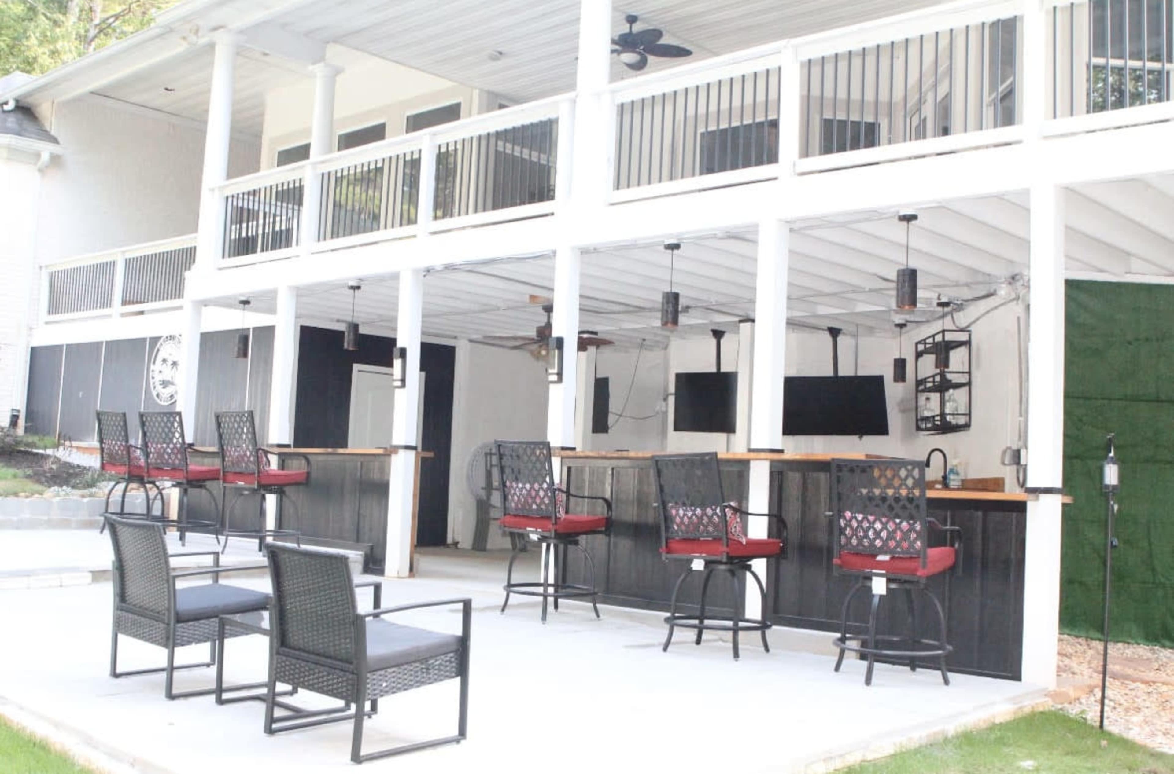 The image shows a spacious outdoor bar area beneath a covered patio, equipped with several high-top chairs and a flat-screen television mounted on the wall.
