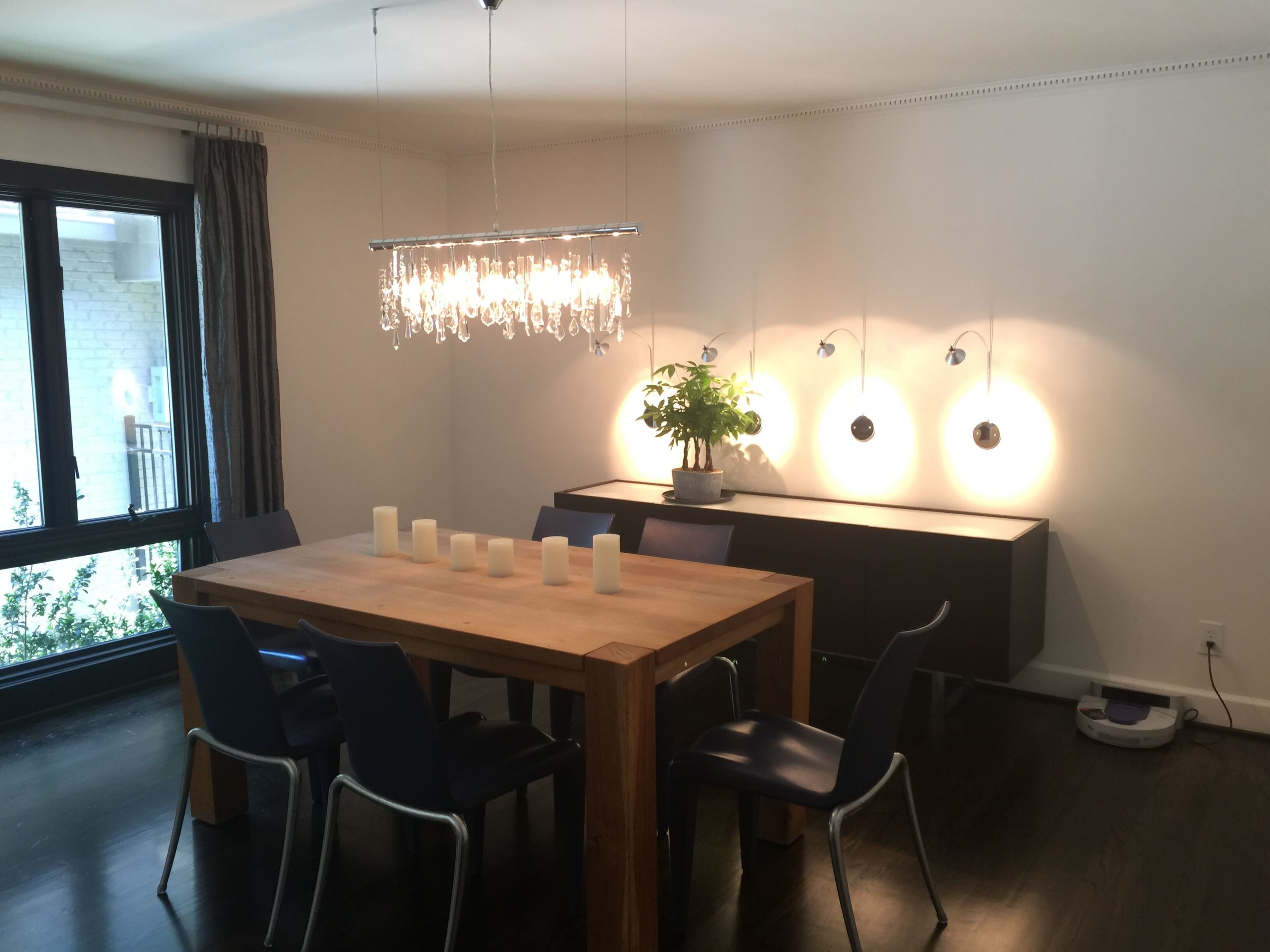 A dining room features a wooden table surrounded by blue chairs, illuminated by a chandelier, with a plant on a sideboard and wall-mounted lights casting circular reflections.