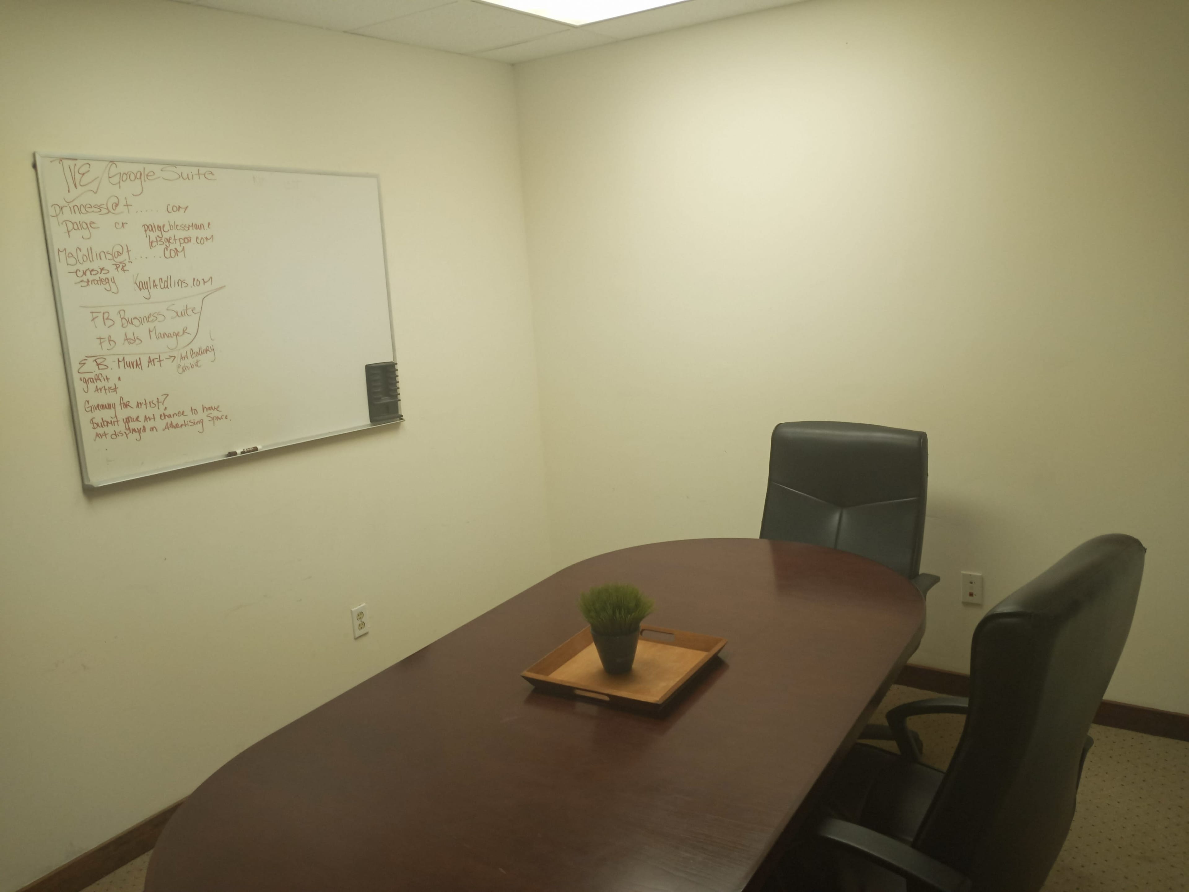A small conference room features a wooden table with a plant centerpiece and two black chairs, alongside a whiteboard displaying handwritten notes.