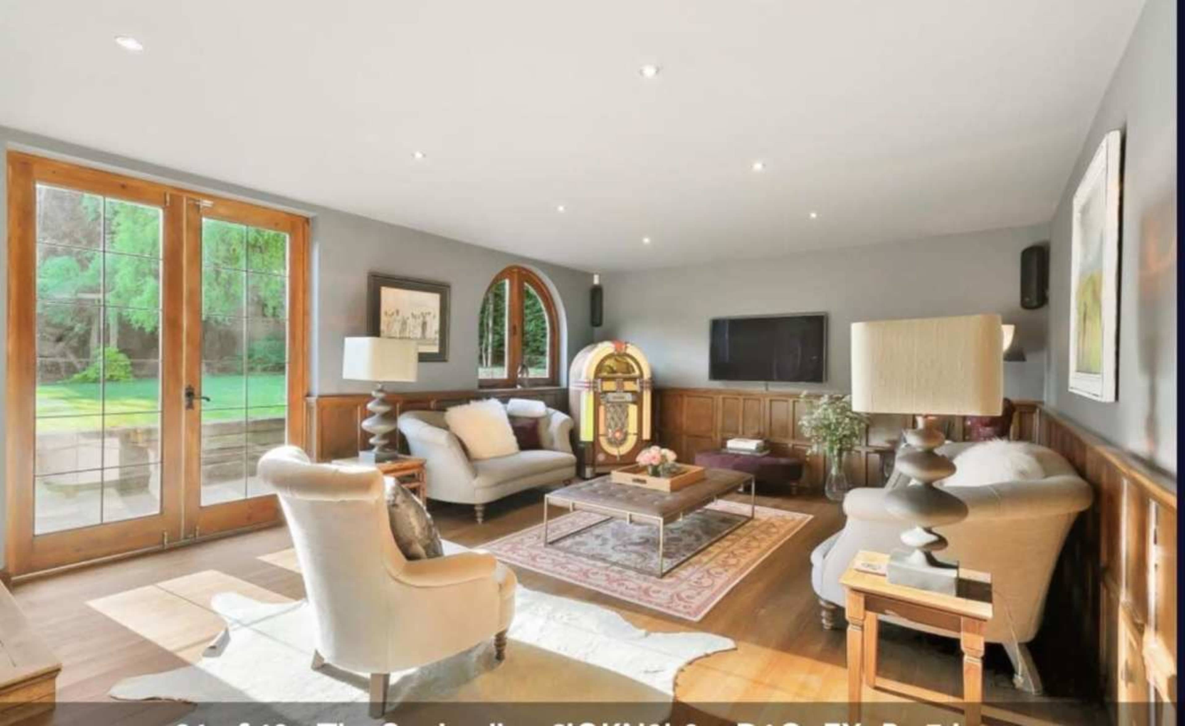 The image shows a well-lit living room featuring two armchairs, a cream-colored sofa, a wooden coffee table, and a vintage jukebox, with large windows that open to a green outdoor area.