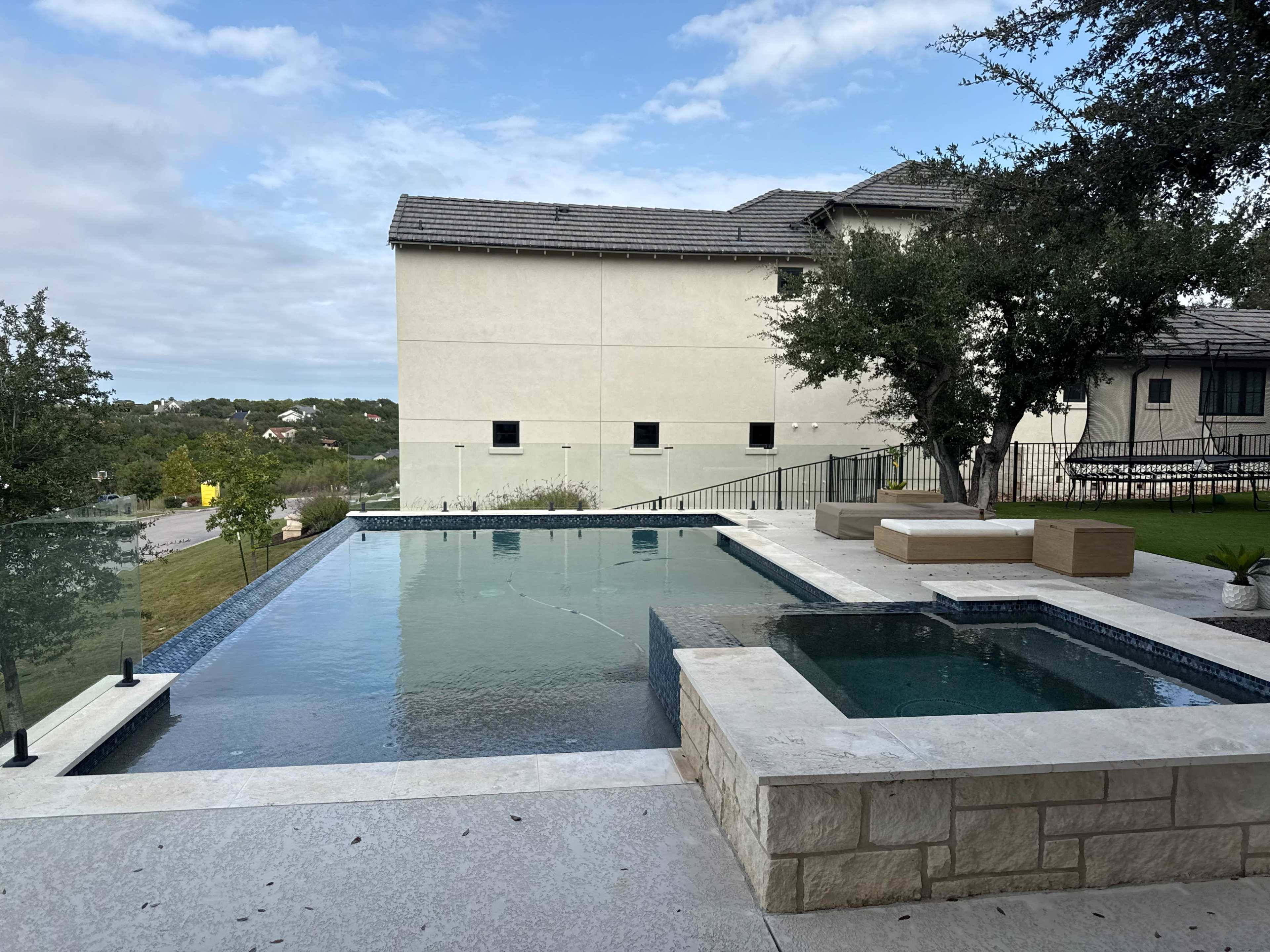 The image shows a modern pool with a spa area, surrounded by stone decking and landscaped greenery, set against a backdrop of a residential building and cloudy sky.