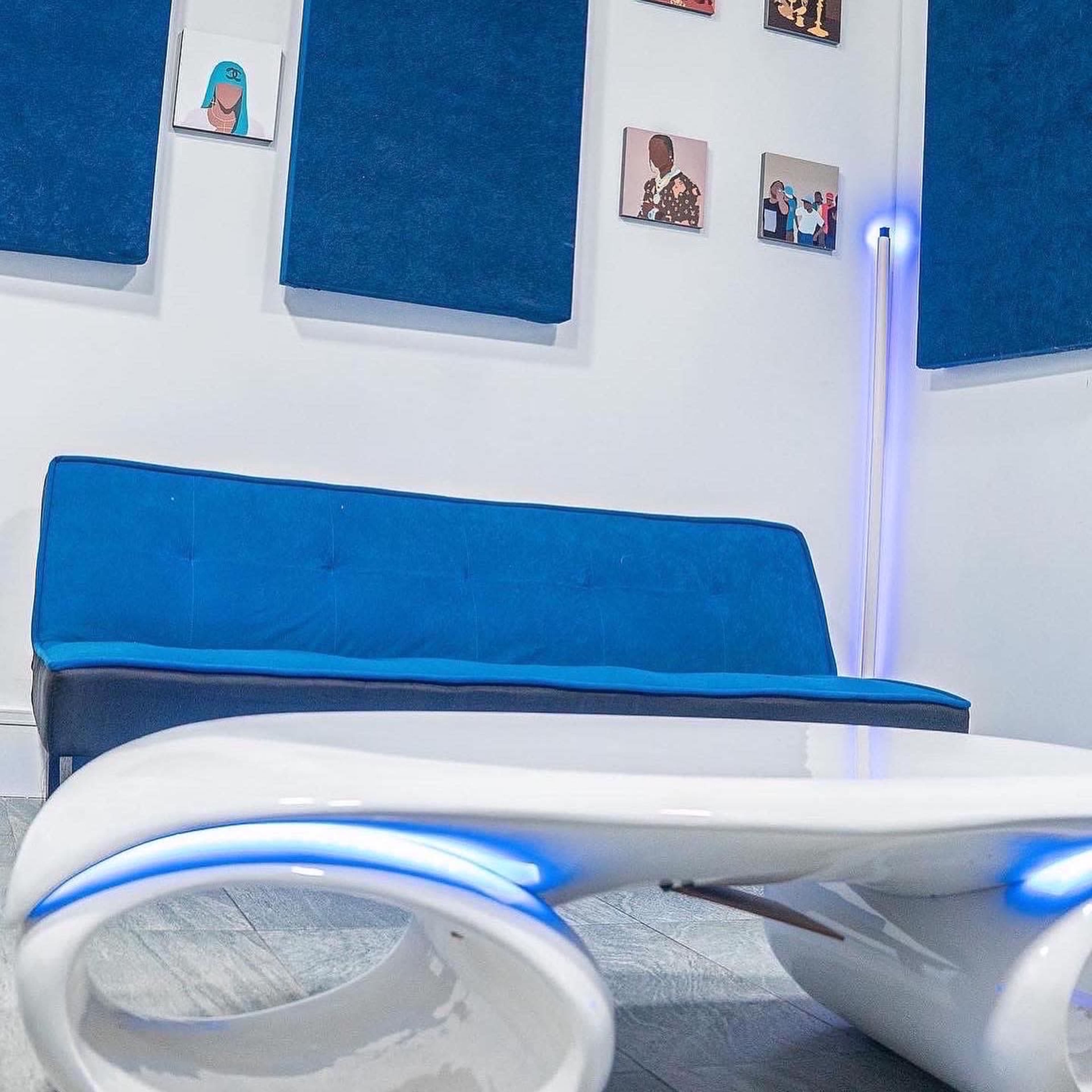 A blue couch in a room with blue wall panels and various framed artworks on the walls, along with a white coffee table featuring blue lighting accents.