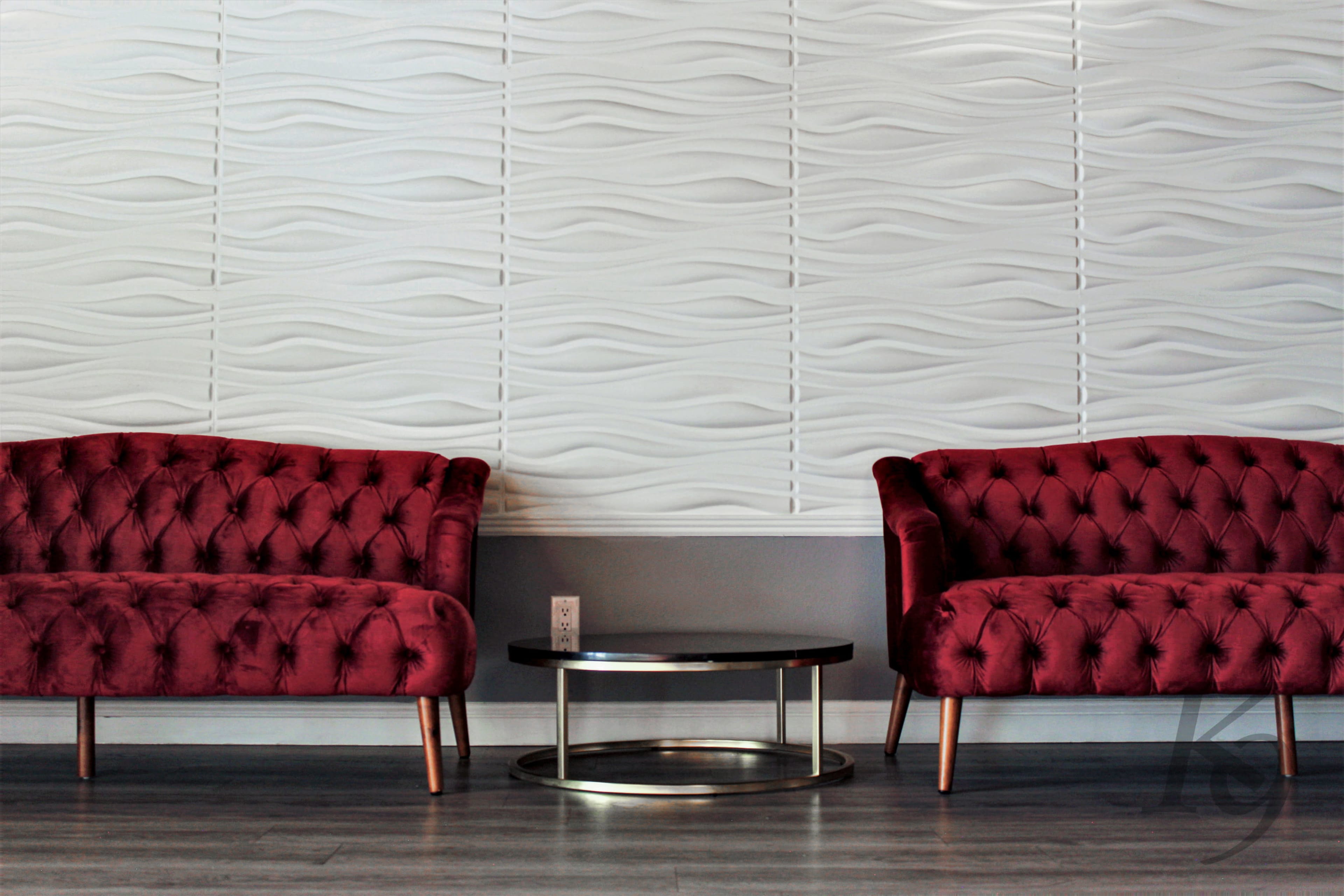 Two red tufted sofas are positioned on either side of a circular metal coffee table, facing a textured white wave-patterned wall.