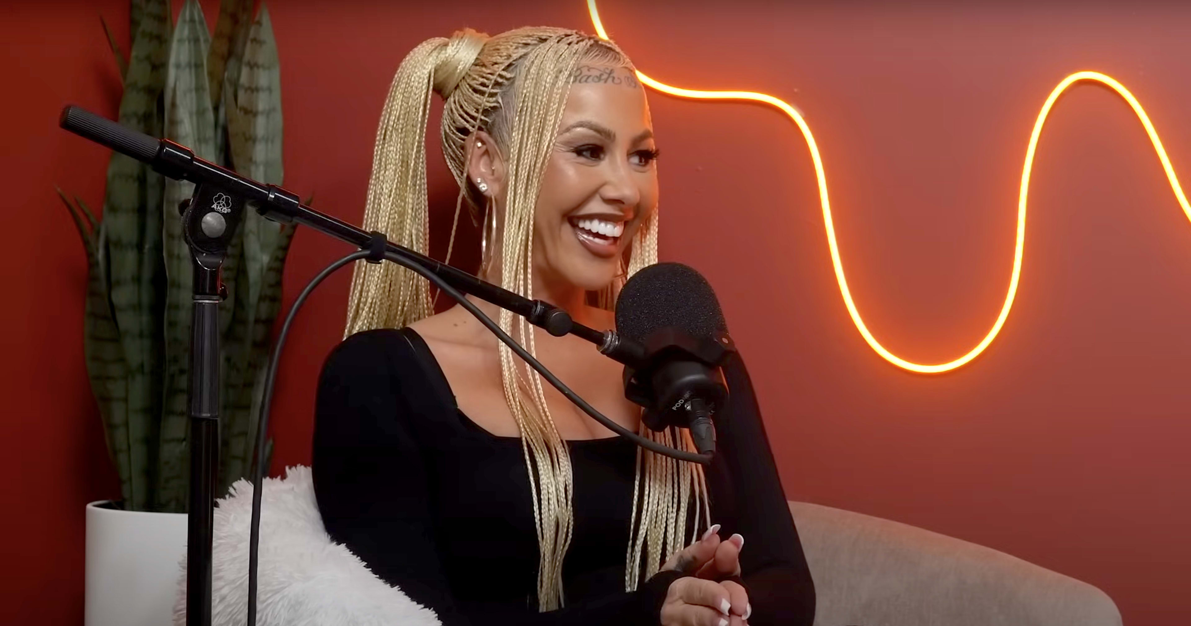 A woman with braided hair smiles while sitting in front of a microphone in a studio with a red wall and a neon light.