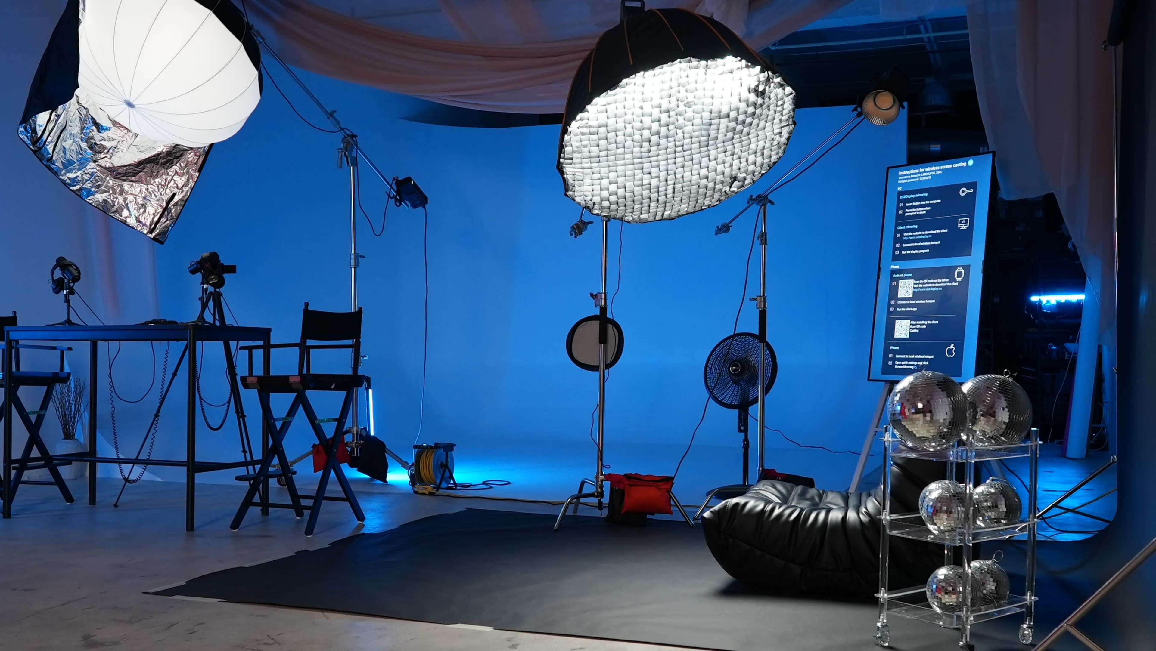 A photography studio setup with lighting equipment, a backdrop, and a display screen, along with chairs and a black beanbag on the floor.