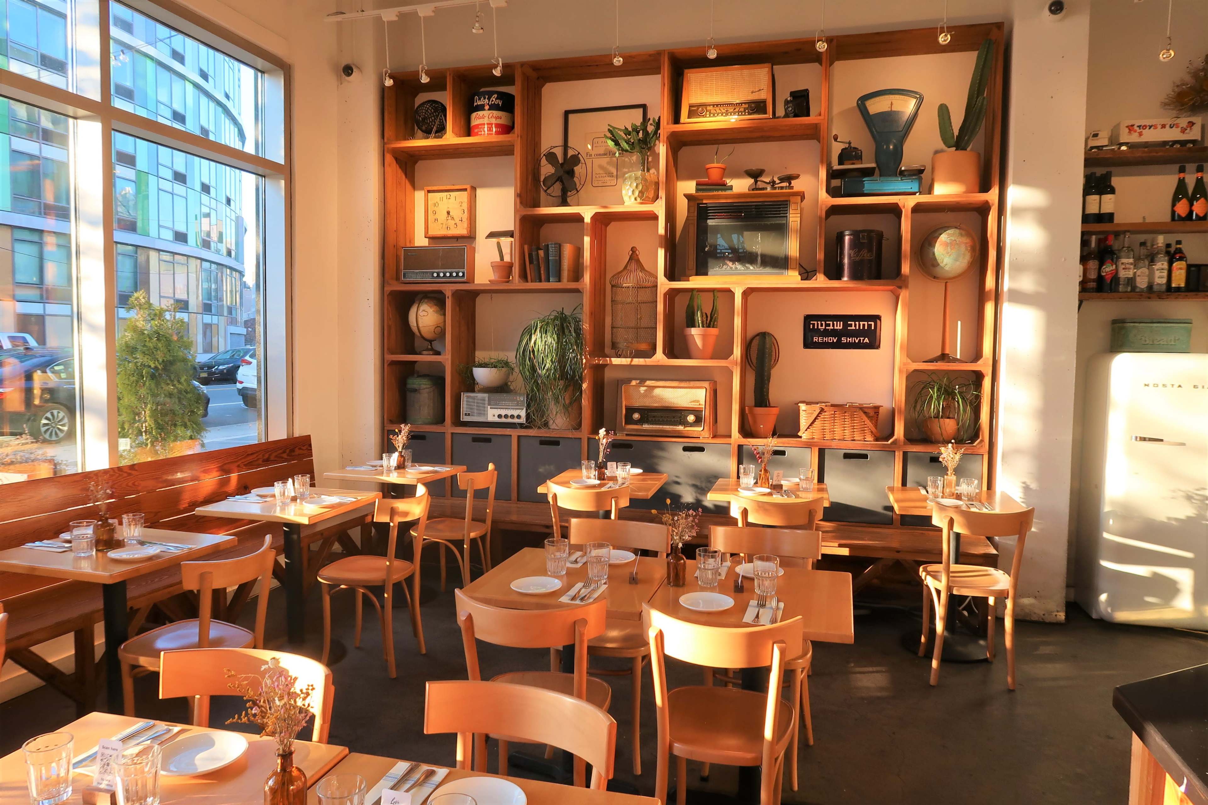 A spacious restaurant features wooden tables set with glassware, surrounded by a decorative wall shelf displaying various vintage items and plants.