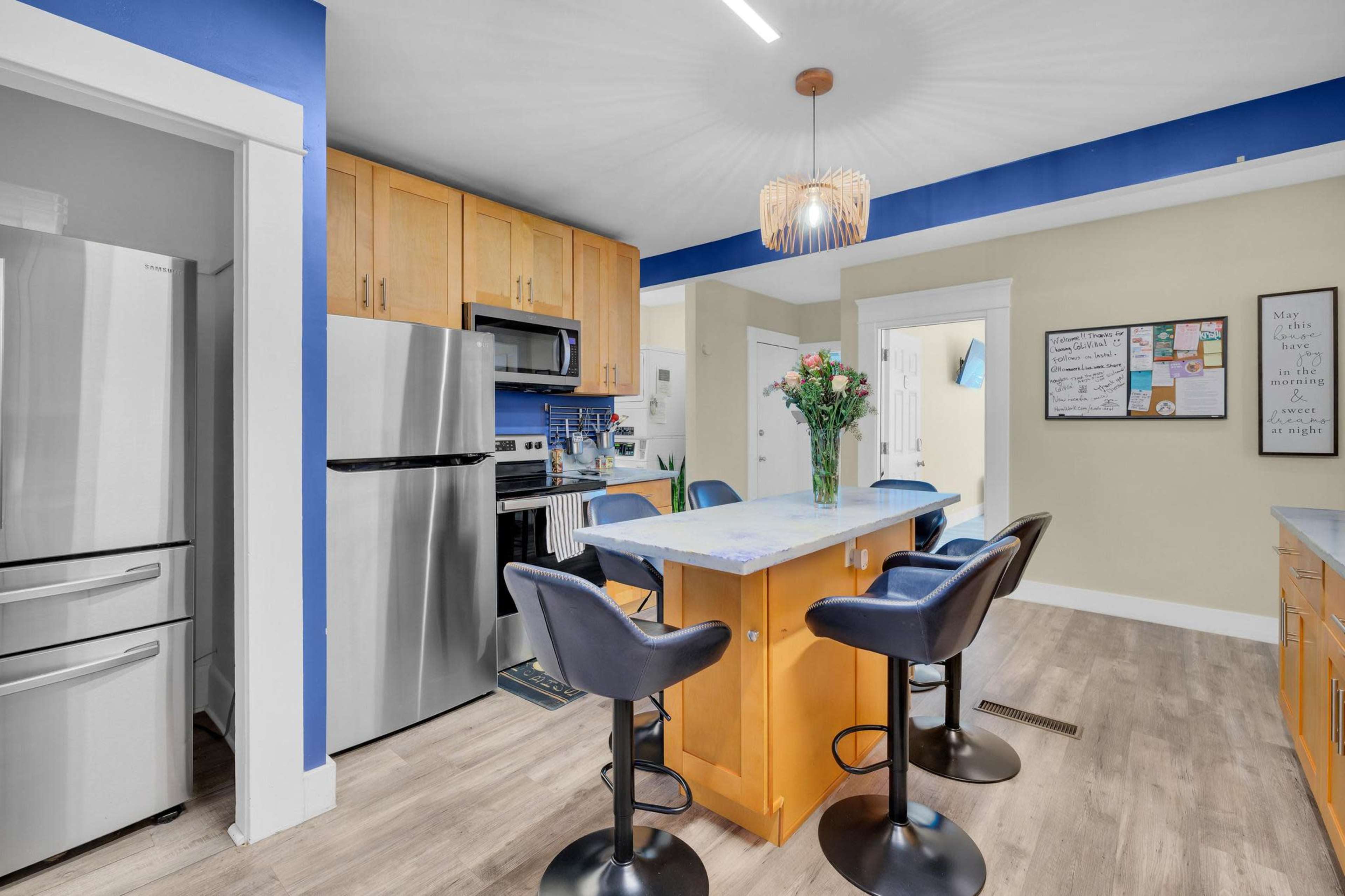A modern kitchen features stainless steel appliances, light wooden cabinets, and a central island with blue seating and a floral centerpiece.