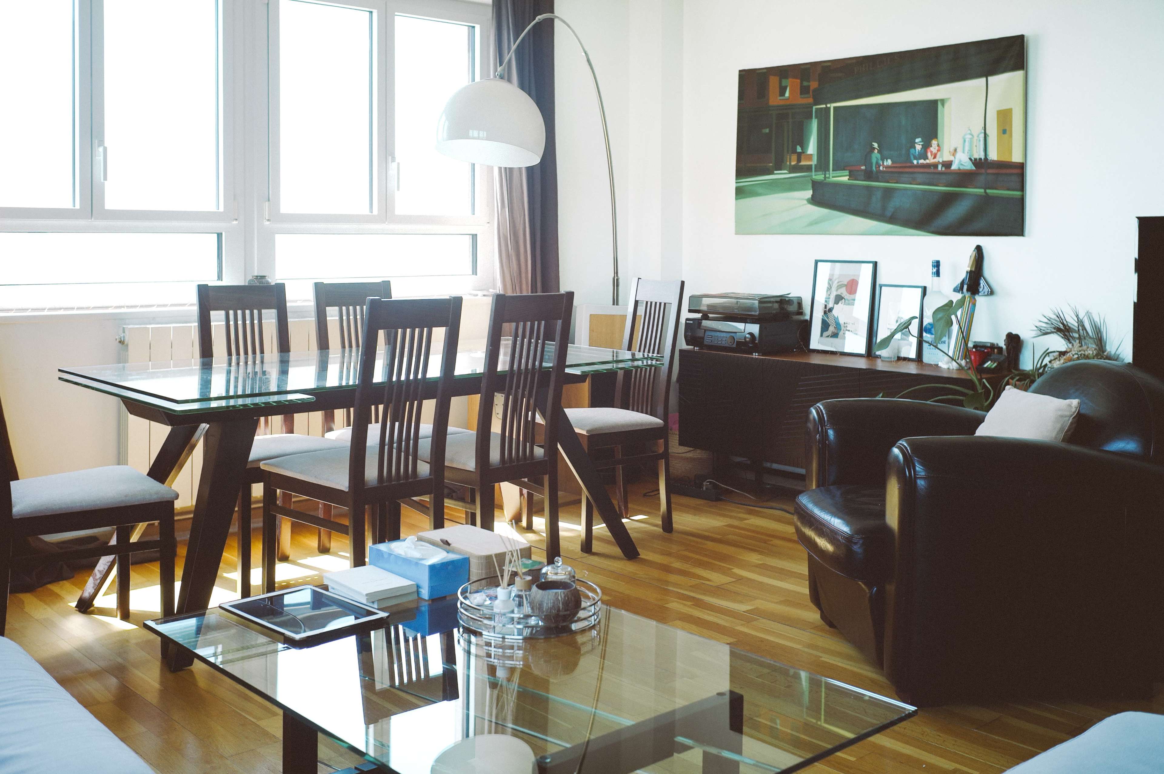 The image shows a modern living room with a glass dining table, six chairs, a black armchair, and a large painting on the wall.