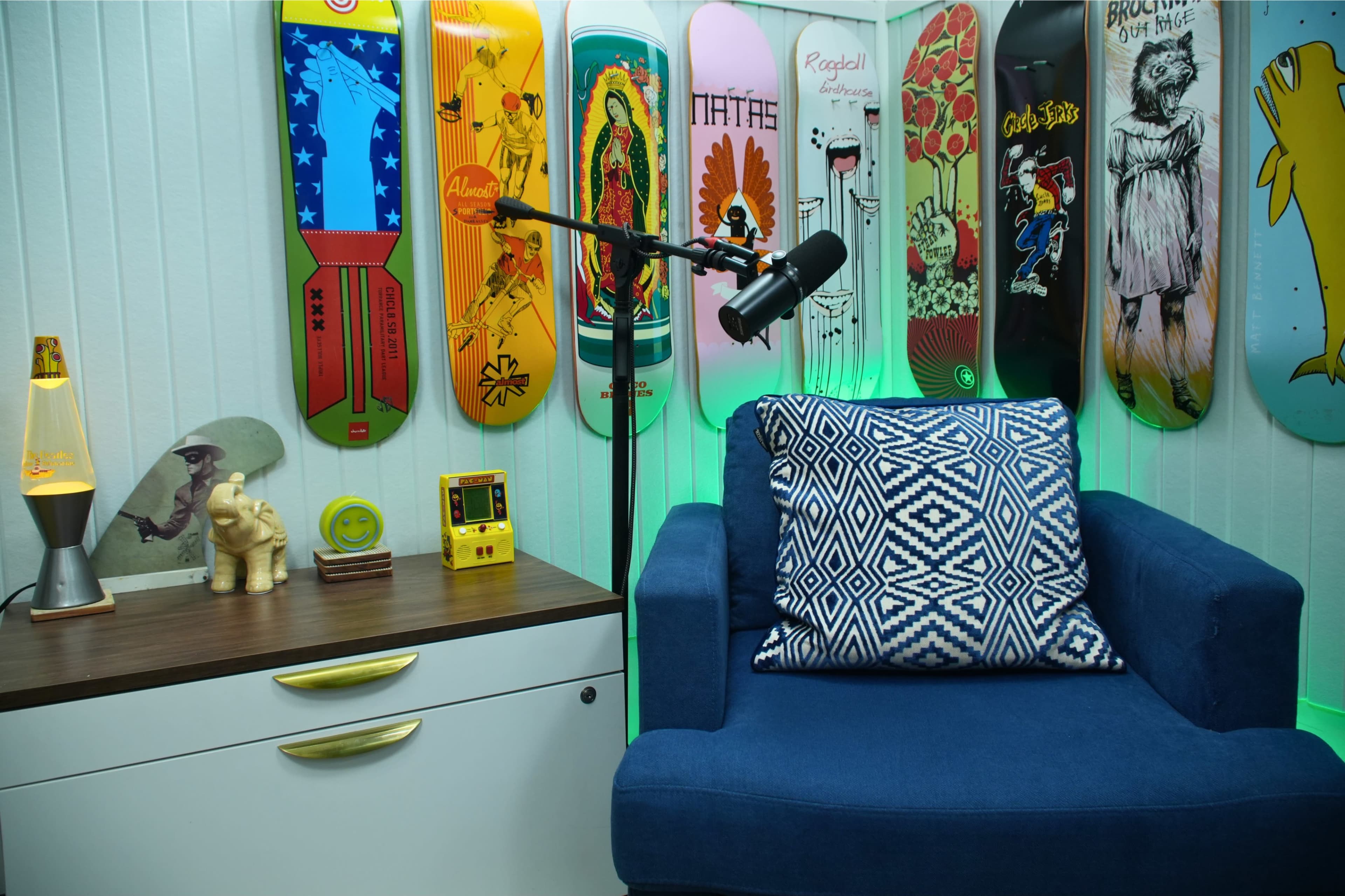 The image shows a cozy corner featuring a blue chair with a patterned pillow, a small table with a lamp and an elephant figurine, and colorful skateboard decks mounted on the wall.