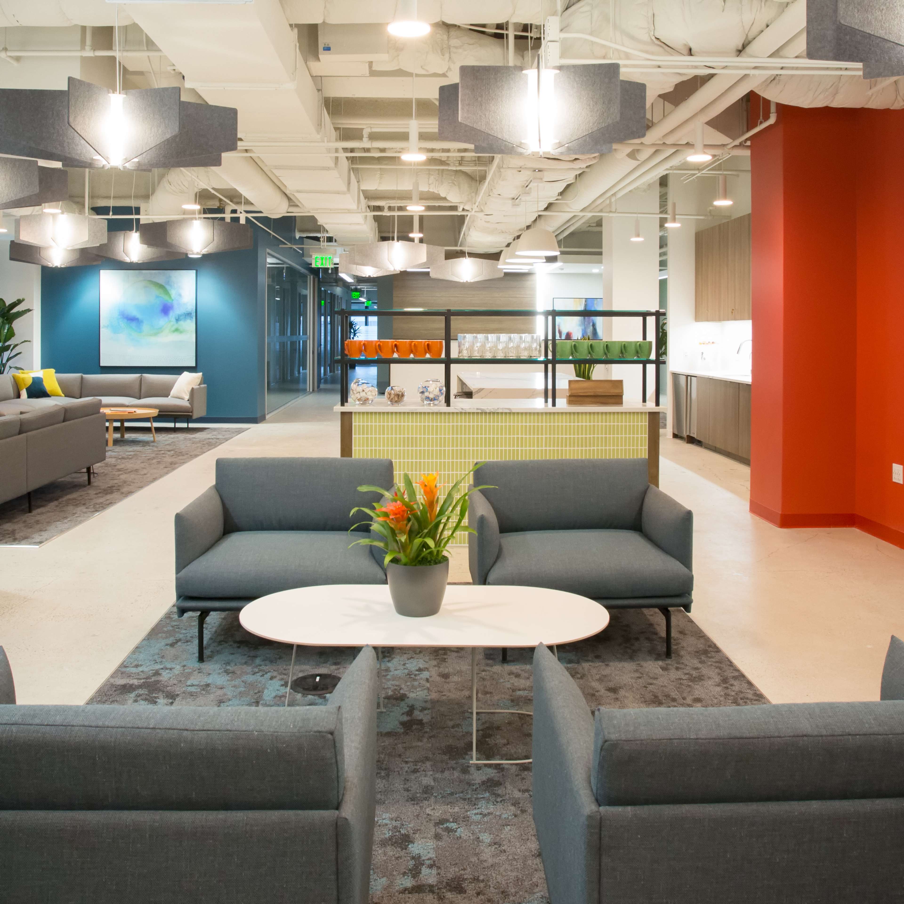 The image shows a modern office lobby area with gray sofas arranged around a round coffee table, decorative plants, and colorful artwork on the walls.