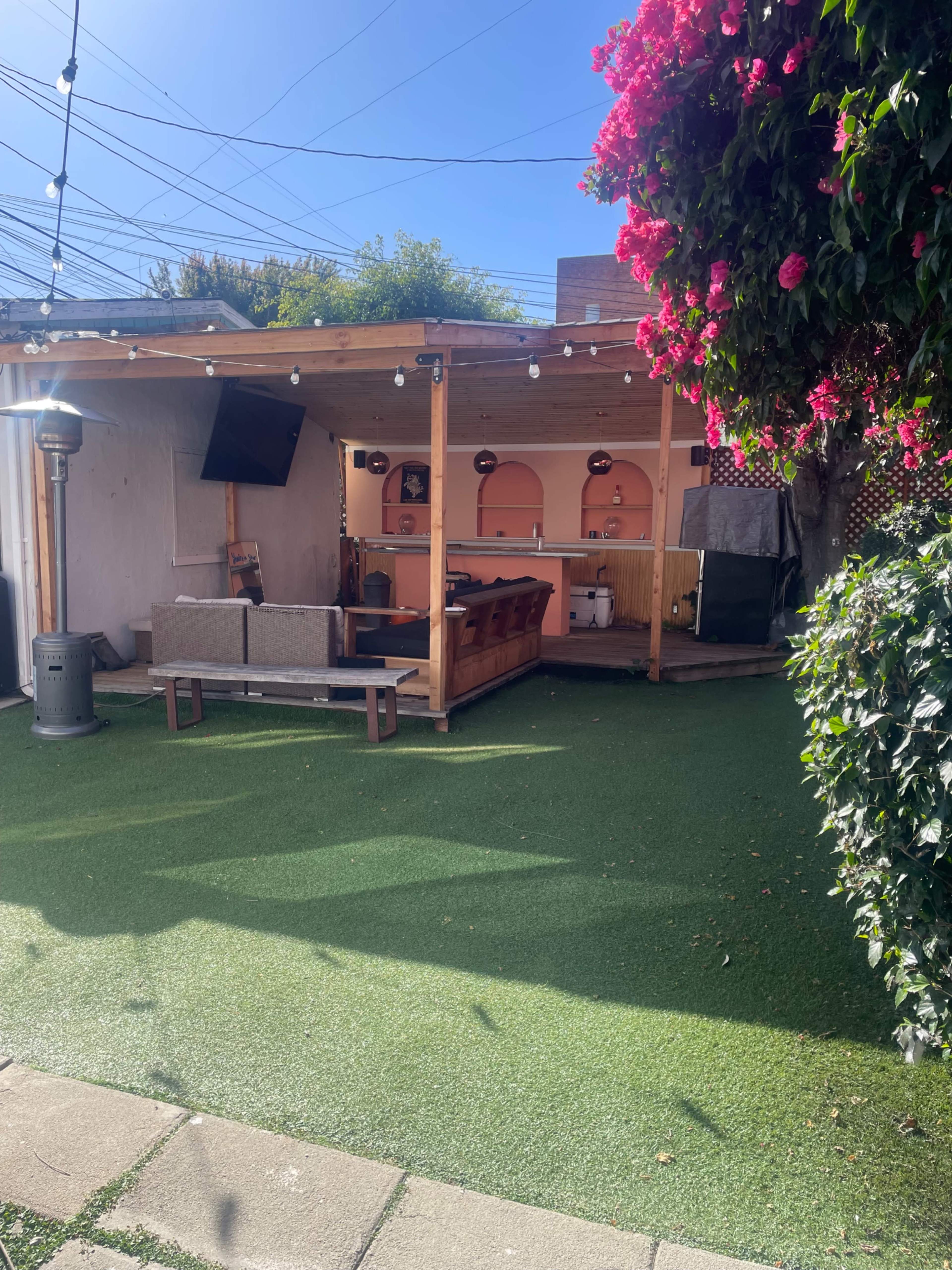 The image shows a backyard area featuring a wooden patio with a couch, a bar setup, and artificial turf, surrounded by greenery and string lights.