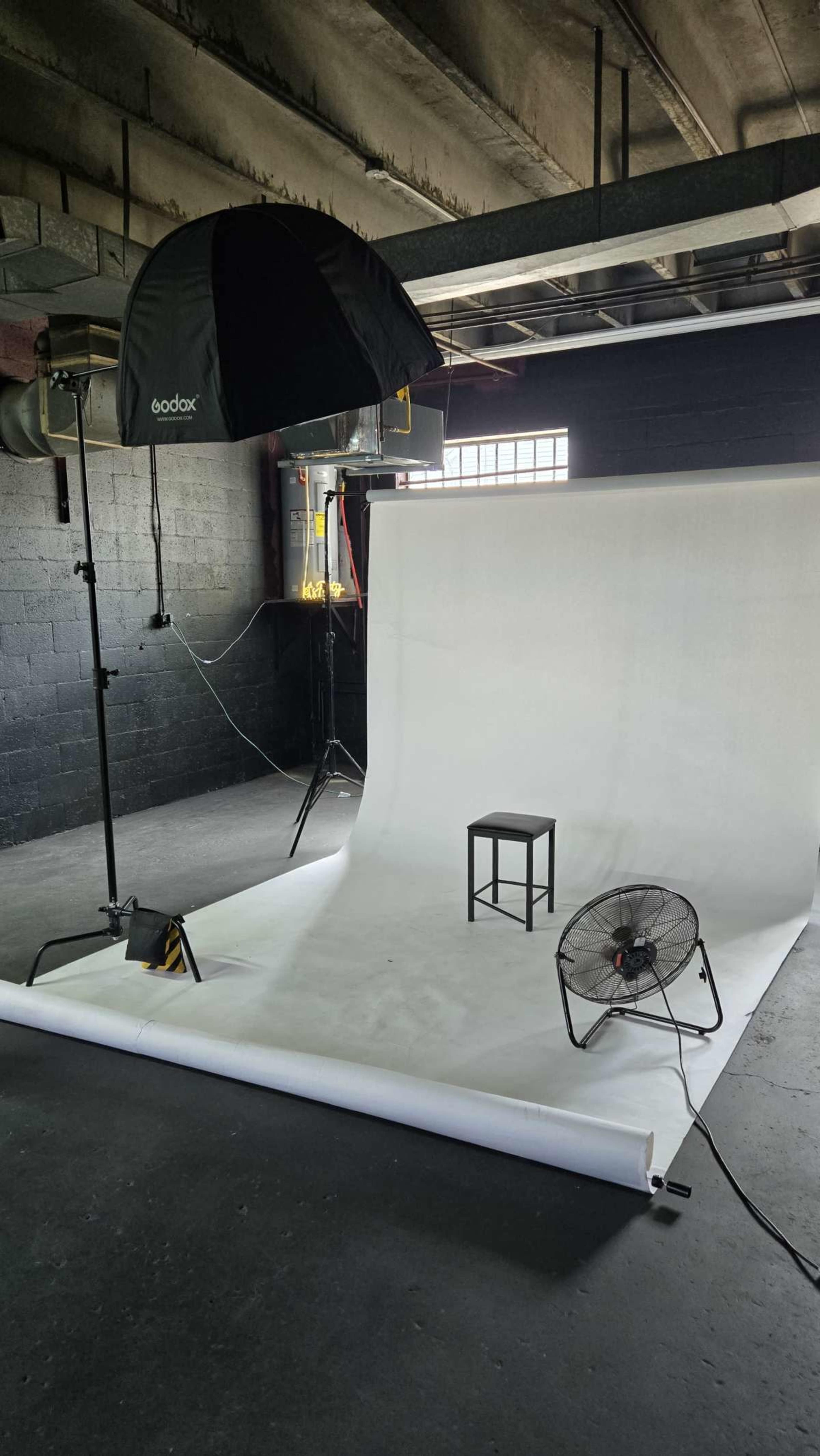The image shows a photography studio setup with a white backdrop, a chair, and a fan.