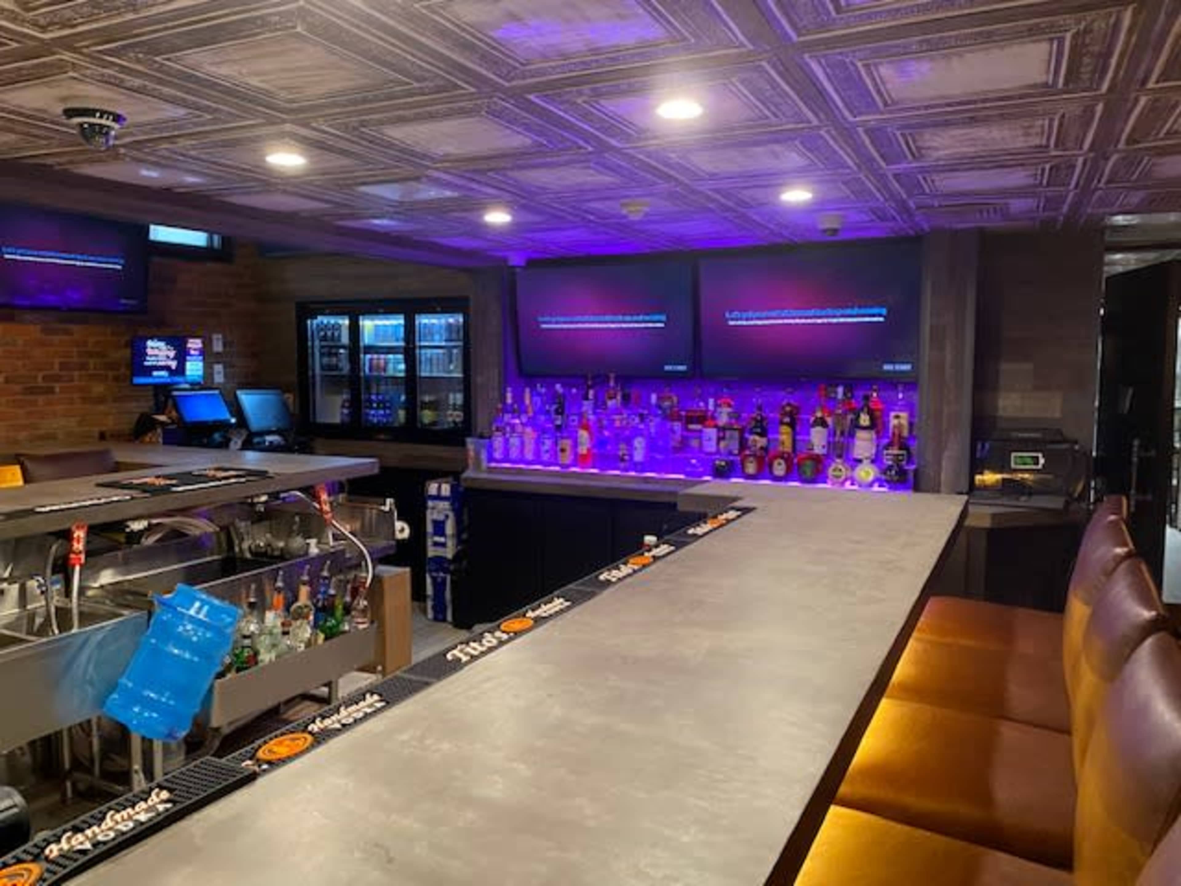 The image shows a bar area with a long countertop, illuminated bottles on shelves, and multiple screens displaying content in a dimly lit setting.