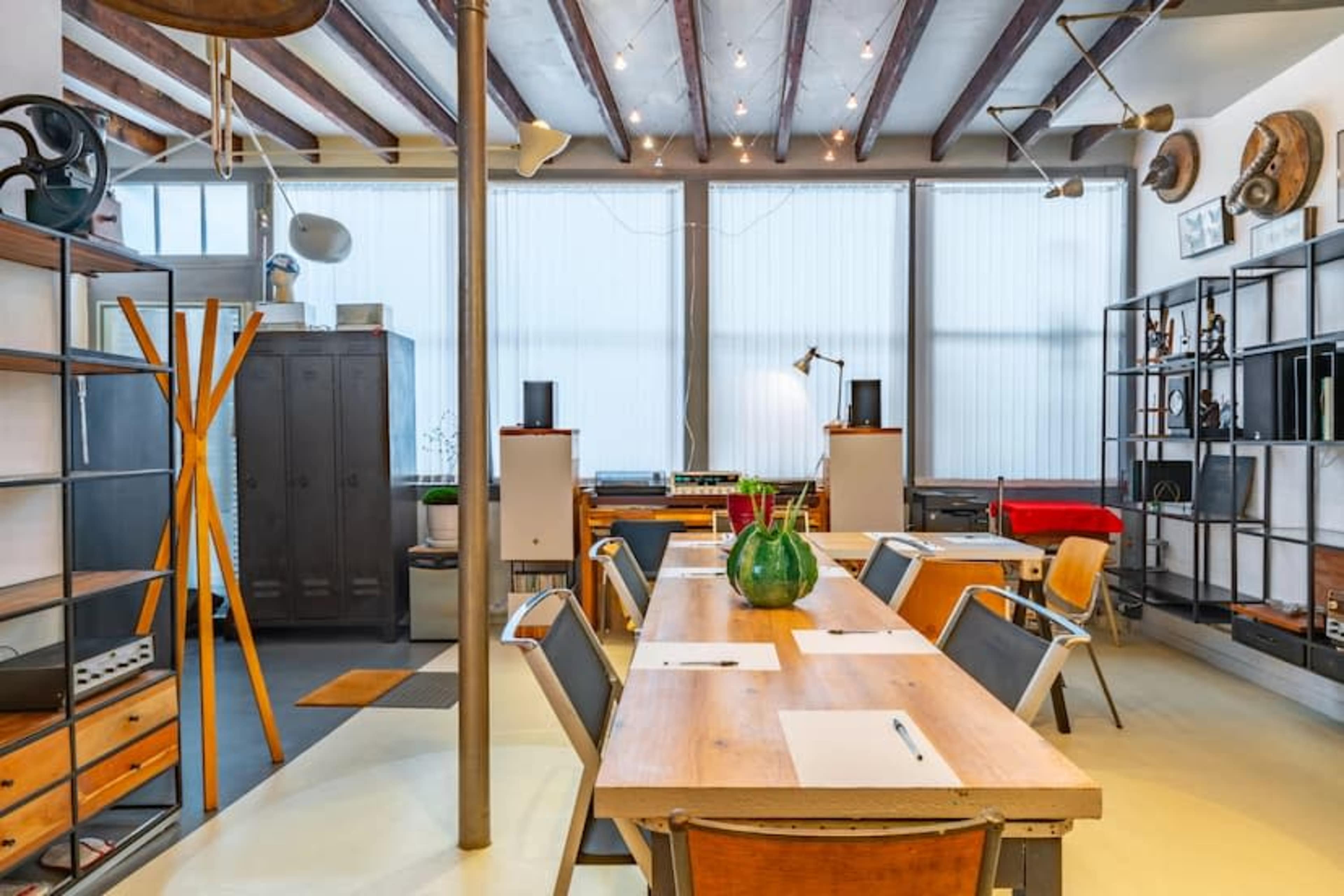 The image shows a modern office space with a long wooden table surrounded by chairs, shelves filled with various items, and large windows providing natural light.