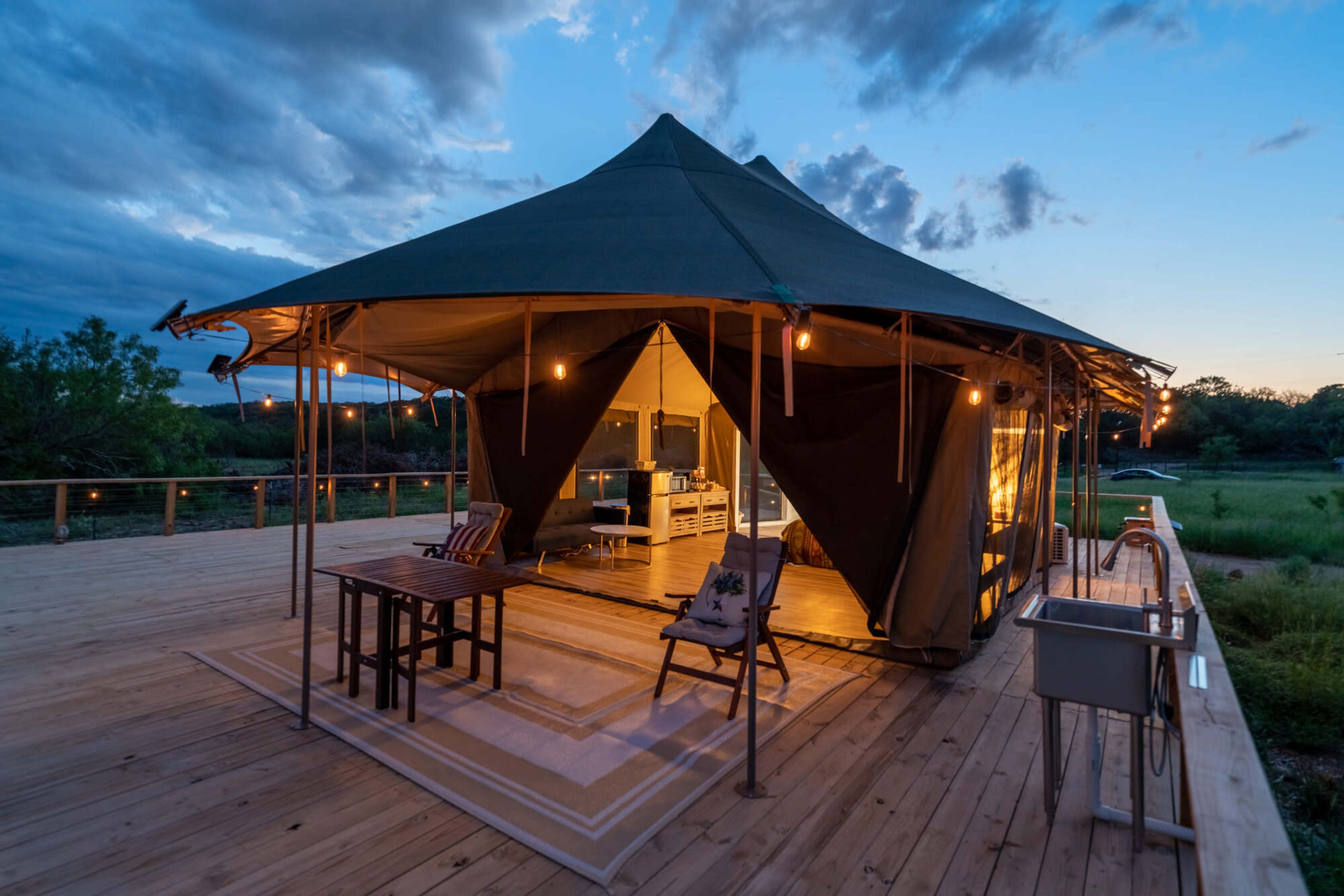 A large canvas tent is set on a wooden deck, with interior furnishings visible and string lights creating ambient lighting at dusk.