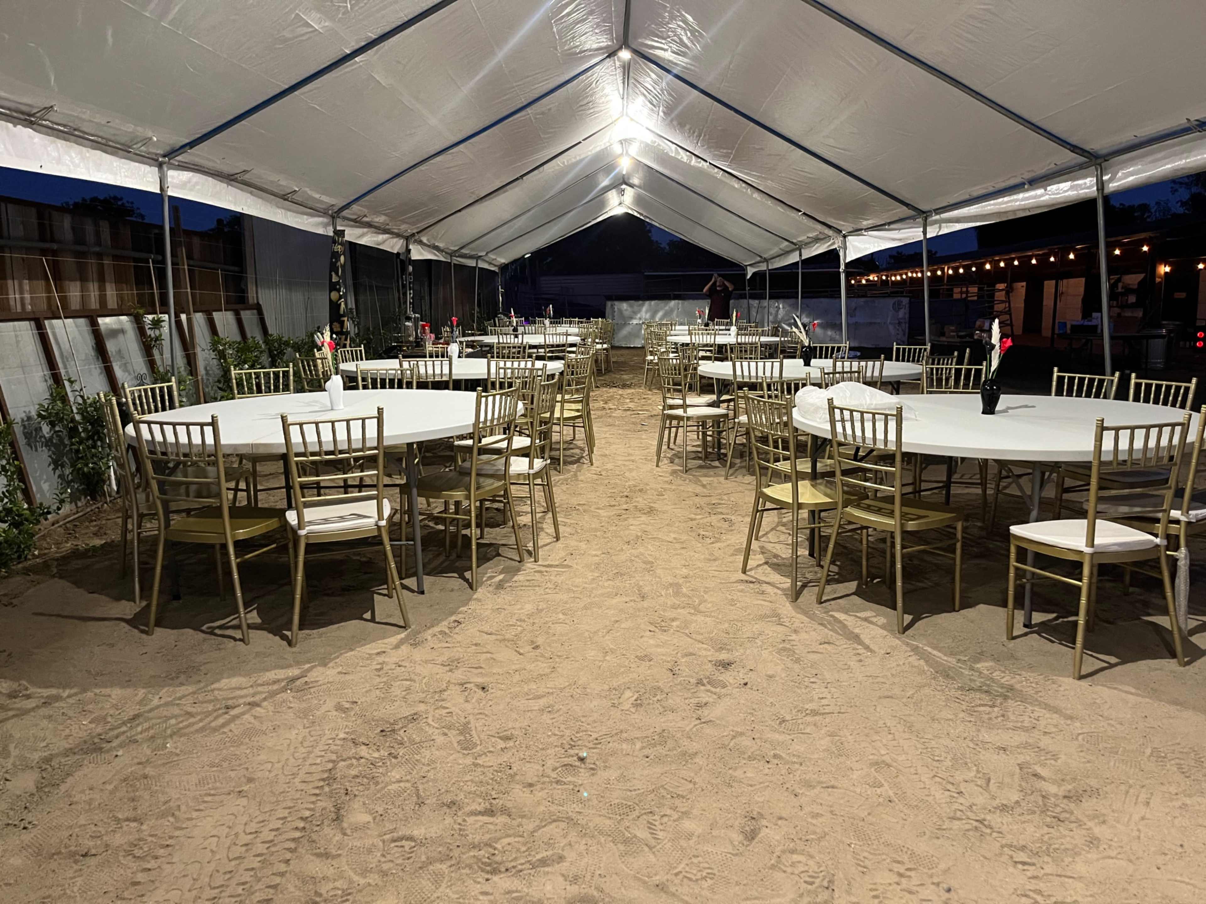A large, open tent is set up with circular tables and chairs arranged on sandy ground, illuminated by overhead lights.