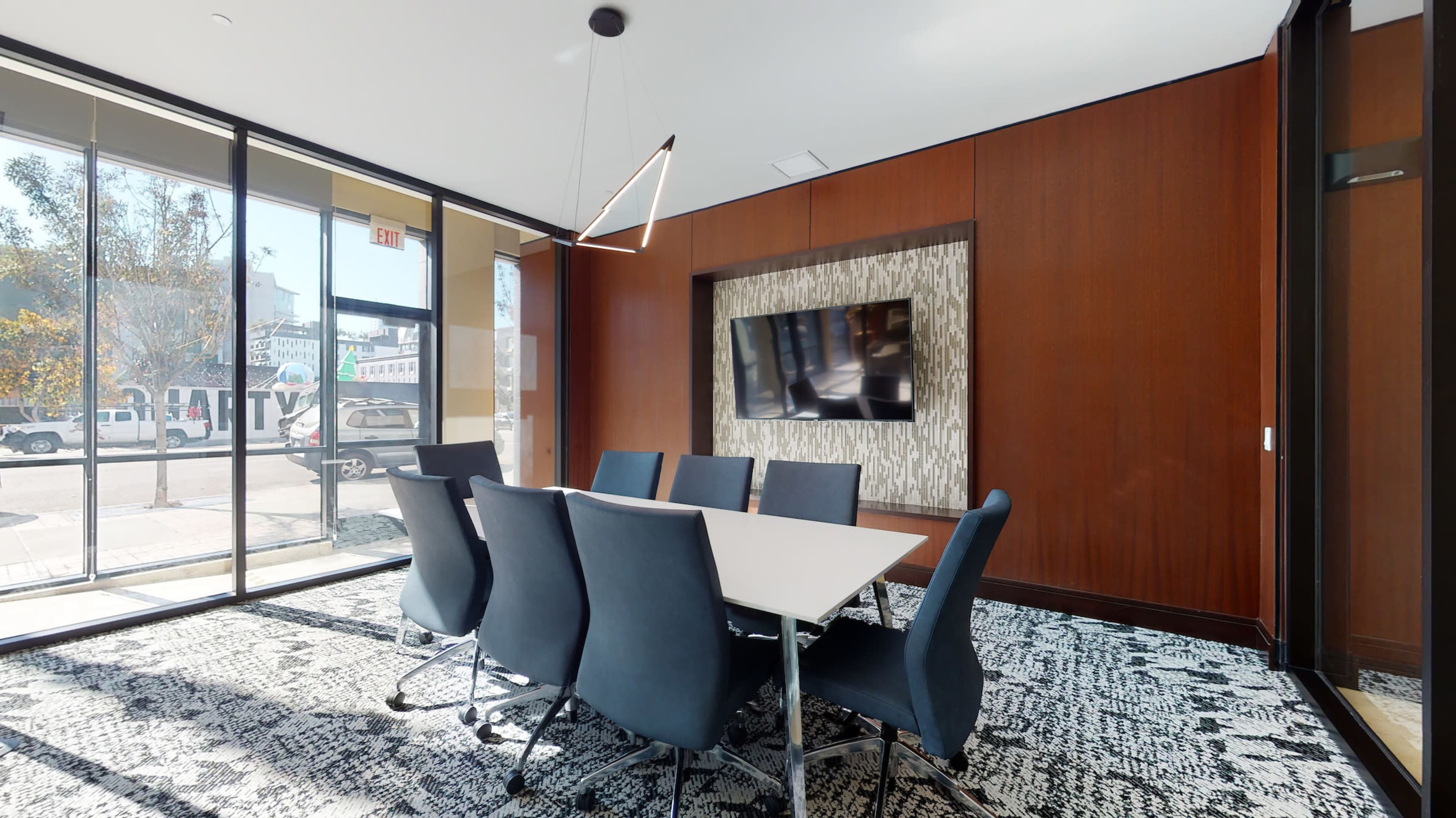 A modern conference room features a long table surrounded by six blue chairs, a large wall-mounted television, and floor-to-ceiling windows allowing natural light to enter.
