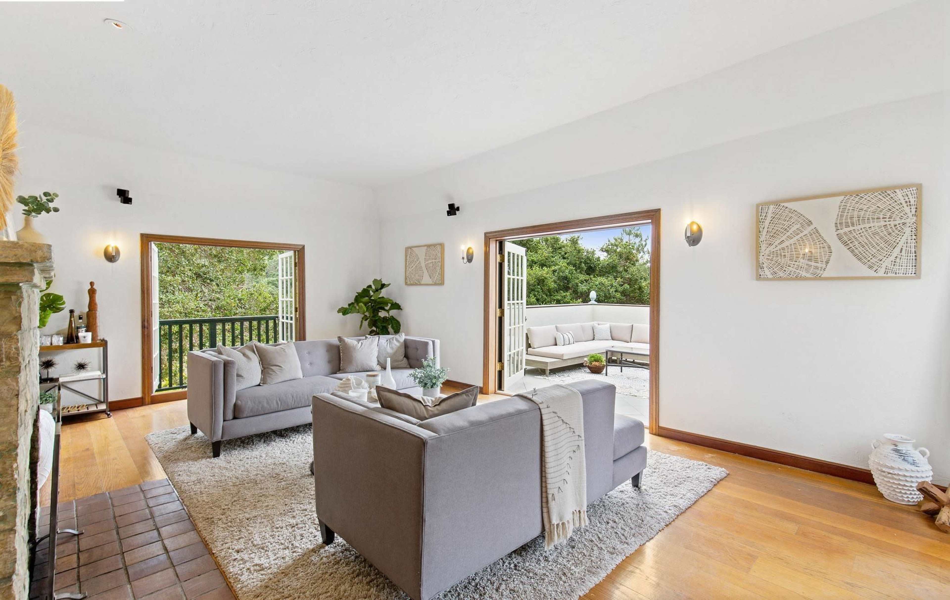 The image shows a bright living room with two gray sofas, a coffee table, and large glass doors leading to an outdoor seating area.