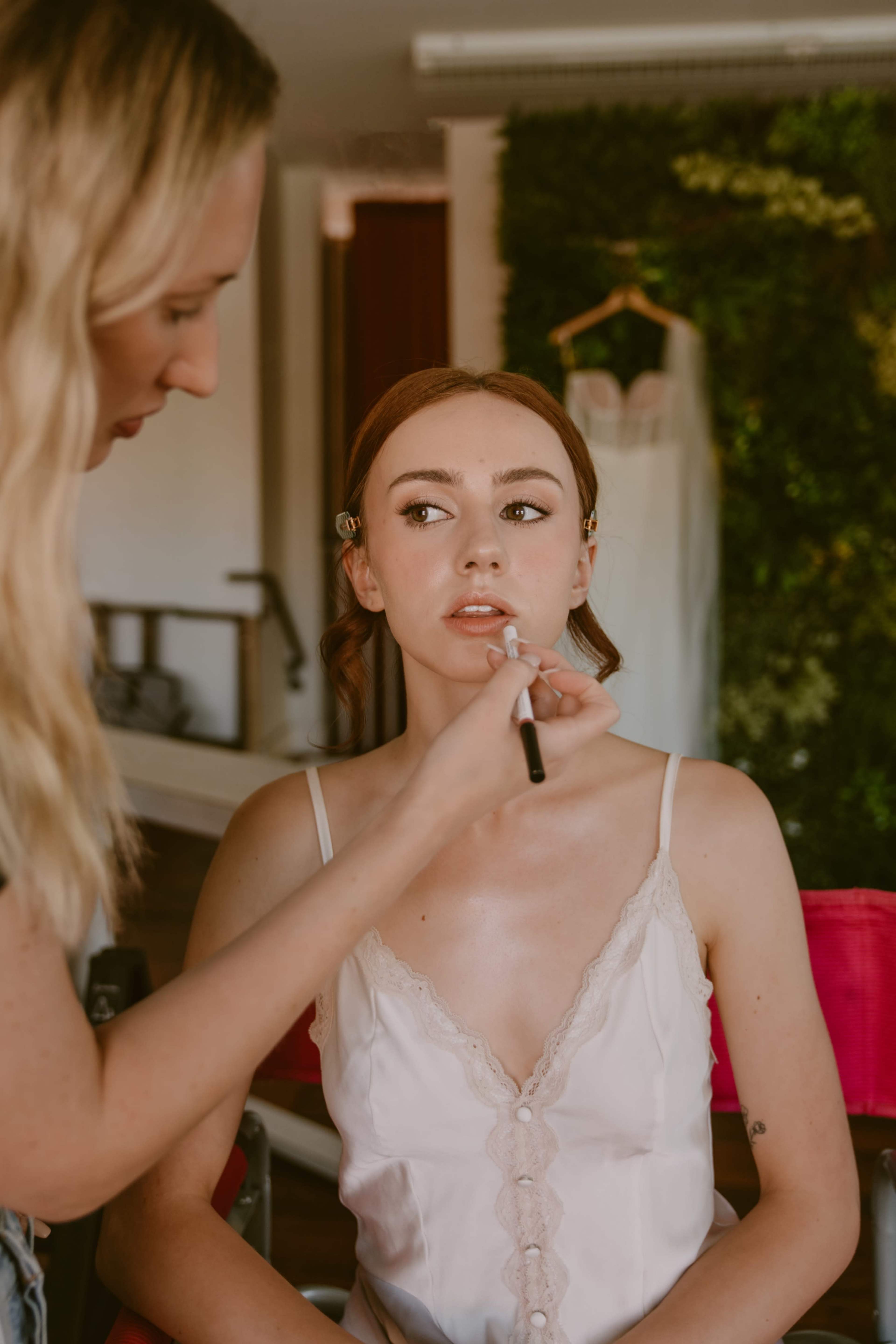 A makeup artist applies lipstick to a woman sitting in a salon chair, with a wedding dress hanging on the wall in the background.