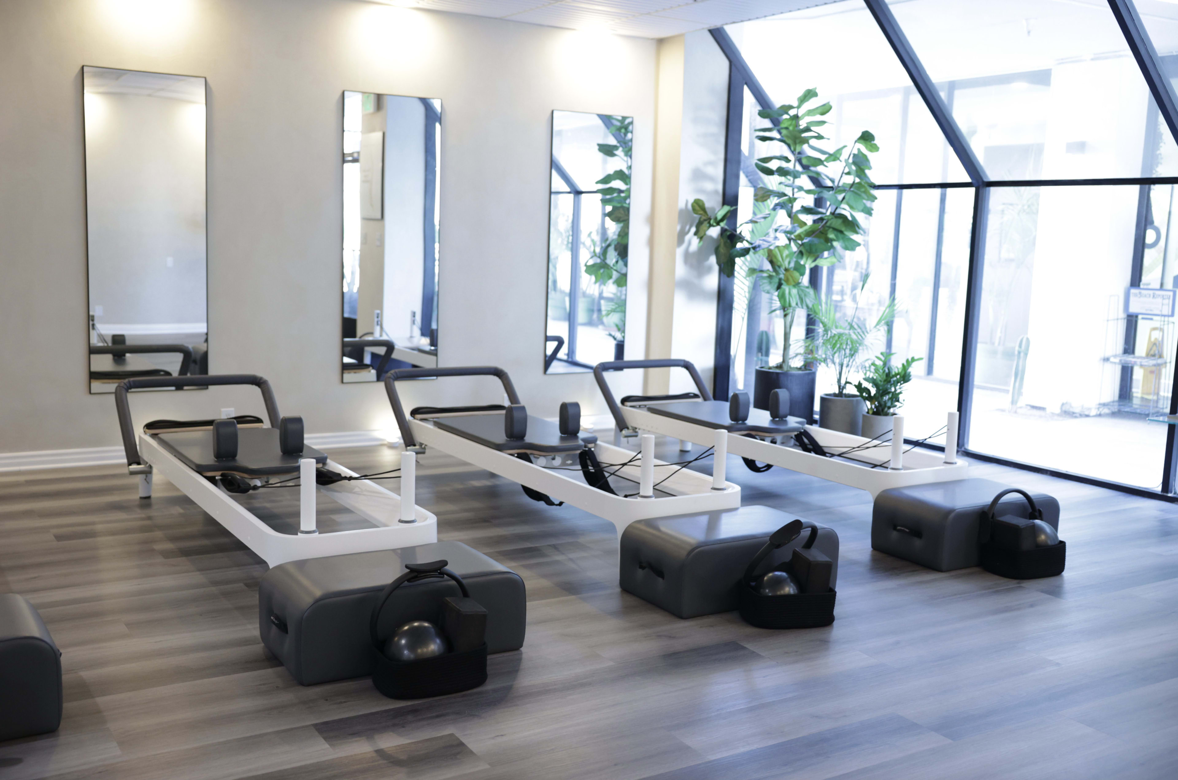 The image shows a modern fitness studio equipped with several reformer Pilates machines arranged neatly along the floor, surrounded by large windows and greenery.