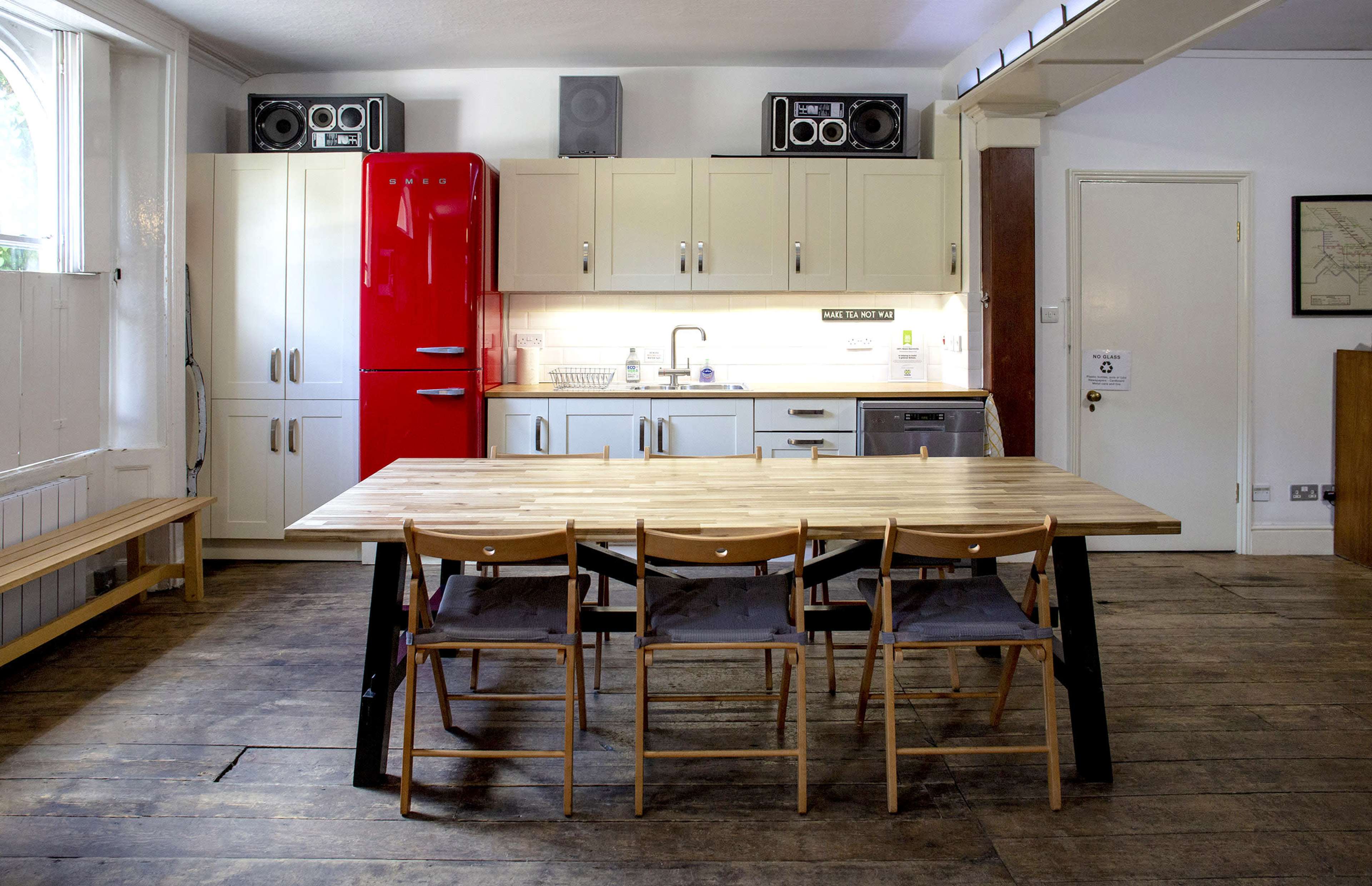 A modern kitchen features a red refrigerator, a wooden dining table with six chairs, and a wall with cabinets and appliances.