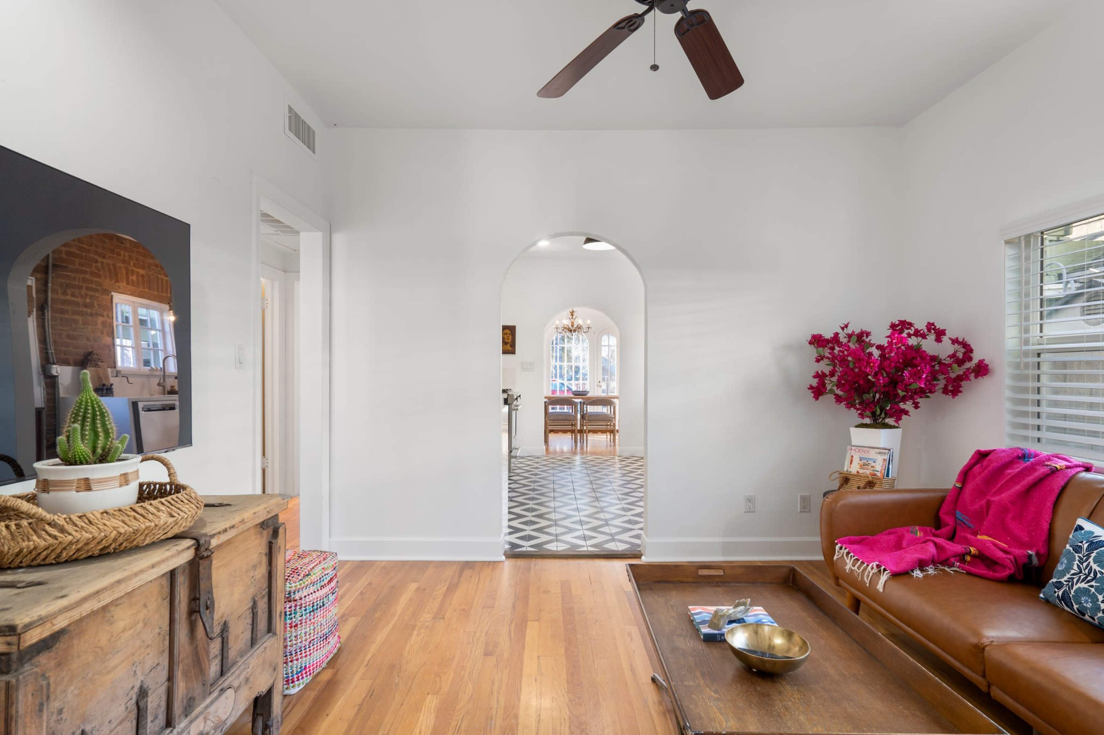 The image shows a bright living room with wooden flooring, a brown leather sofa, and an archway leading to a dining area with a patterned floor.